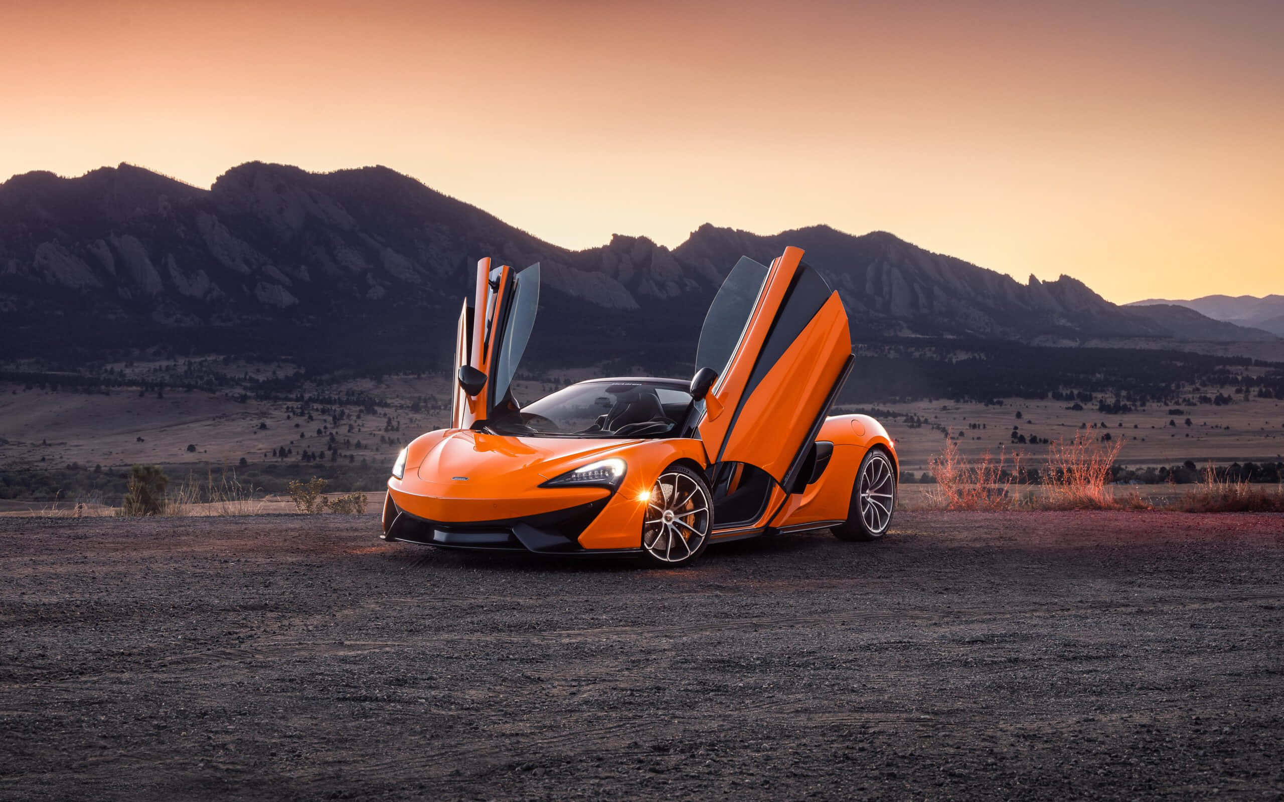 麥拿侖 桌布 壁紙 — 一架充滿活力的橙色McLaren跑車，鷗翼車門打開，停在夕陽下的礫石平原上，背景是崎嶇的山脈。