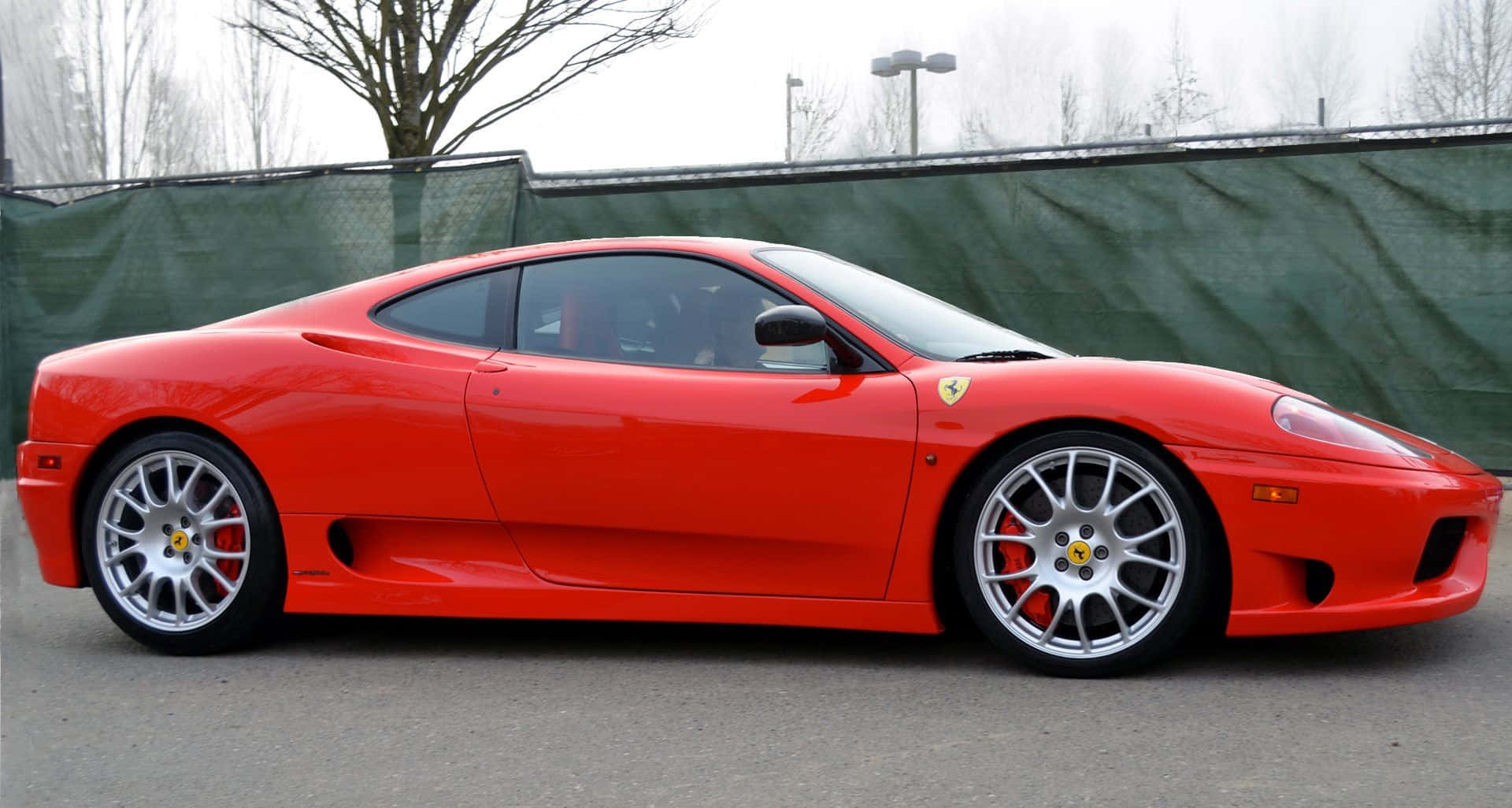 Ferrari 360 Modena 桌布 壁紙 — 一架充滿活力的紅色法拉利360跑車停泊在人行道上，背景有光禿禿的樹木和綠色圍欄。