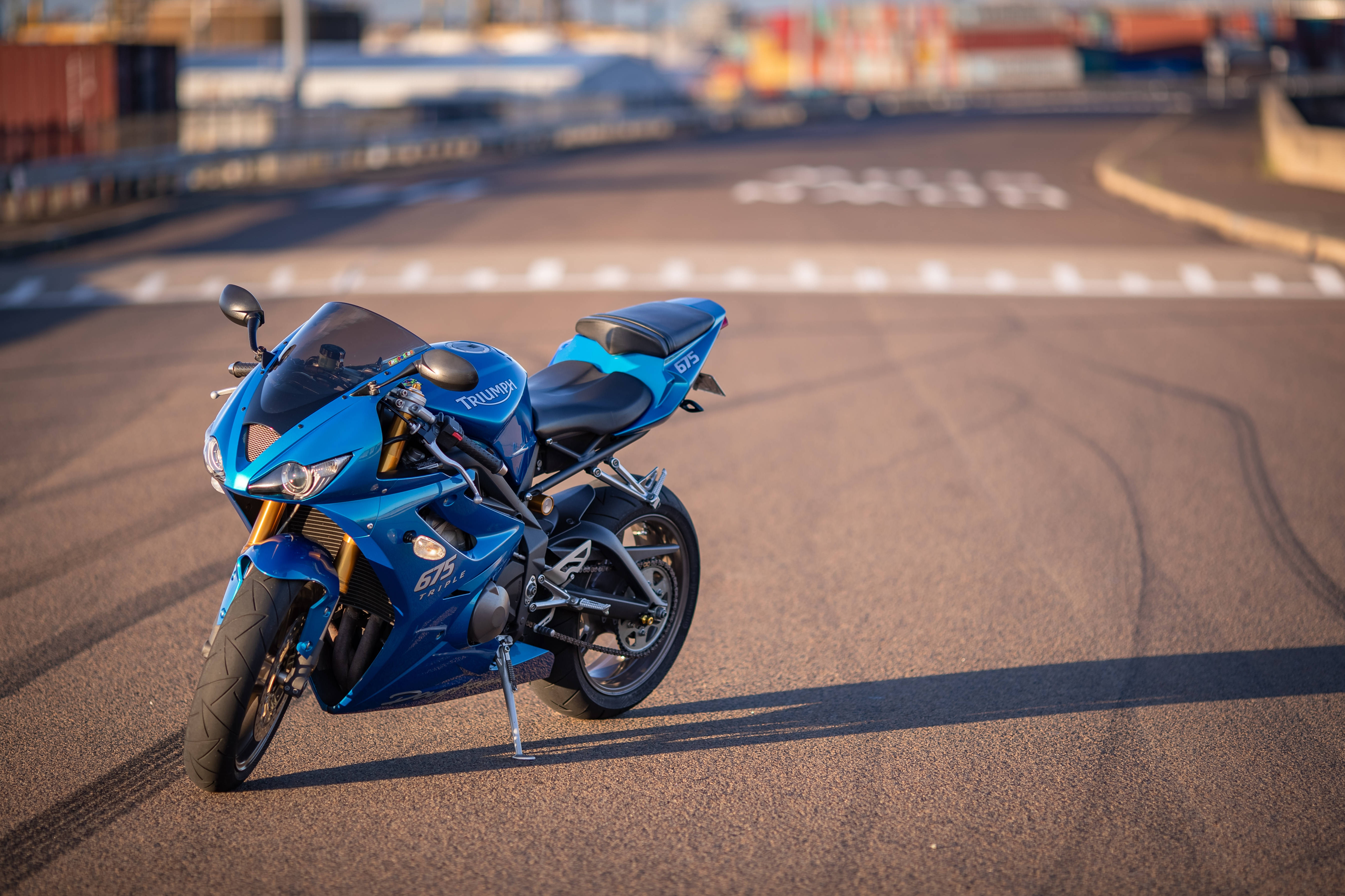 機車 桌布 壁紙 — 一架充滿活力的藍色 Triumph 電單車停泊在陽光普照的賽道上，背景有輪胎痕跡和模糊的貨運集裝箱。