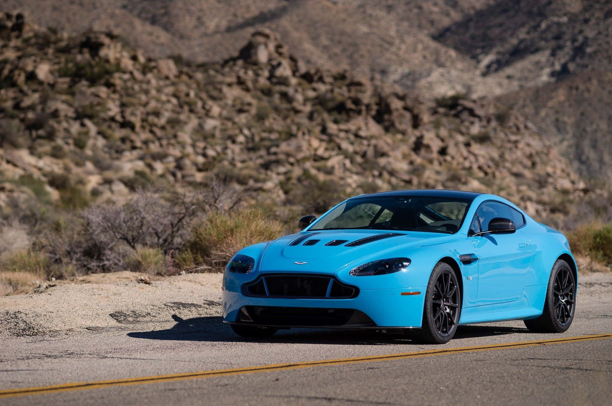 Aston Martin V12 Vantage 桌布 壁紙 — 一架充滿活力的藍色Aston Martin跑車停泊在沙漠公路上，背景是岩石丘陵。