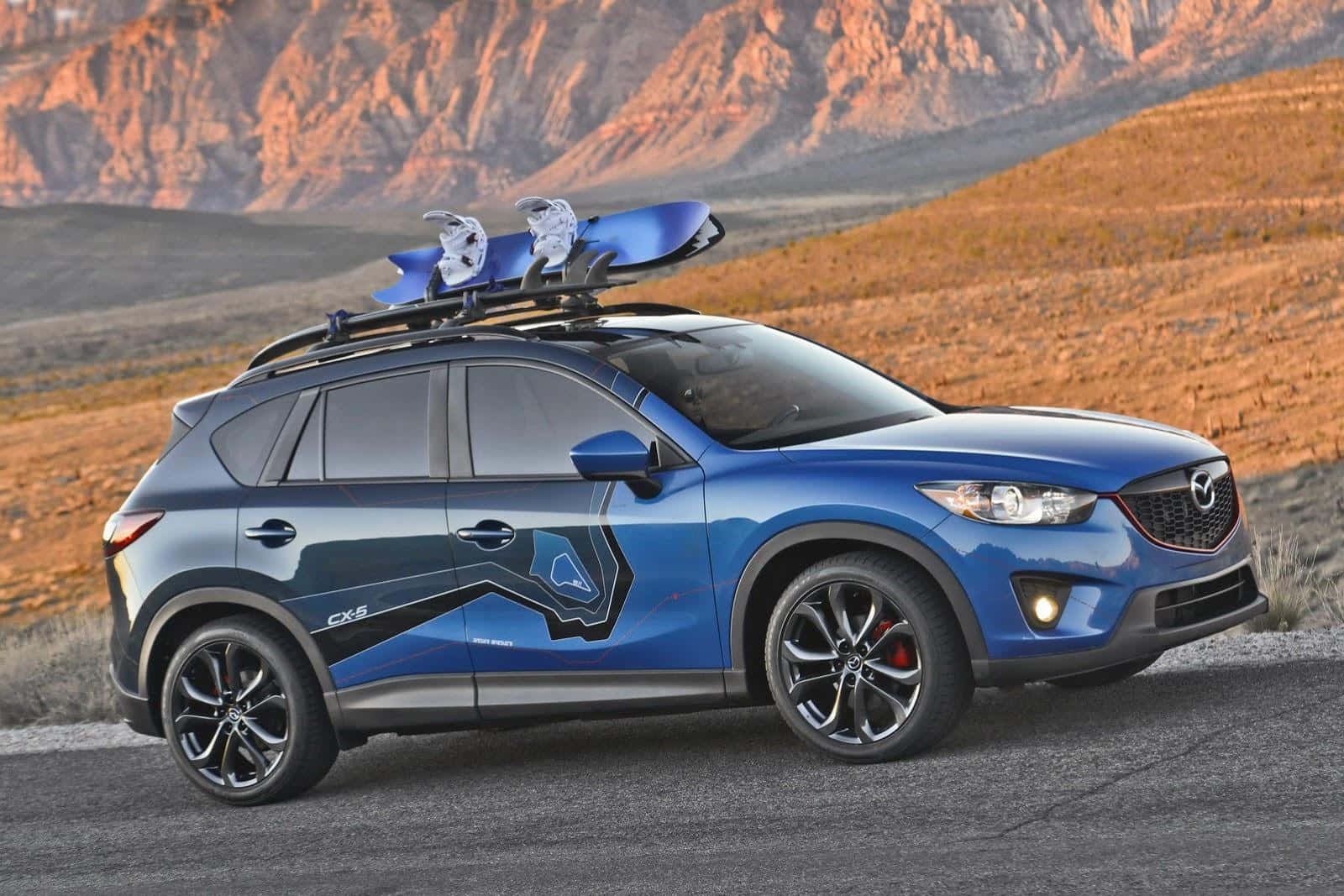 Mazda Cx-5 桌布 壁紙 — 一架充滿活力的藍色Mazda CX-5 SUV，車頂架上放著滑雪板，停在沙漠公路上，背景是崎嶇的橙色山脈，正值日落時分。