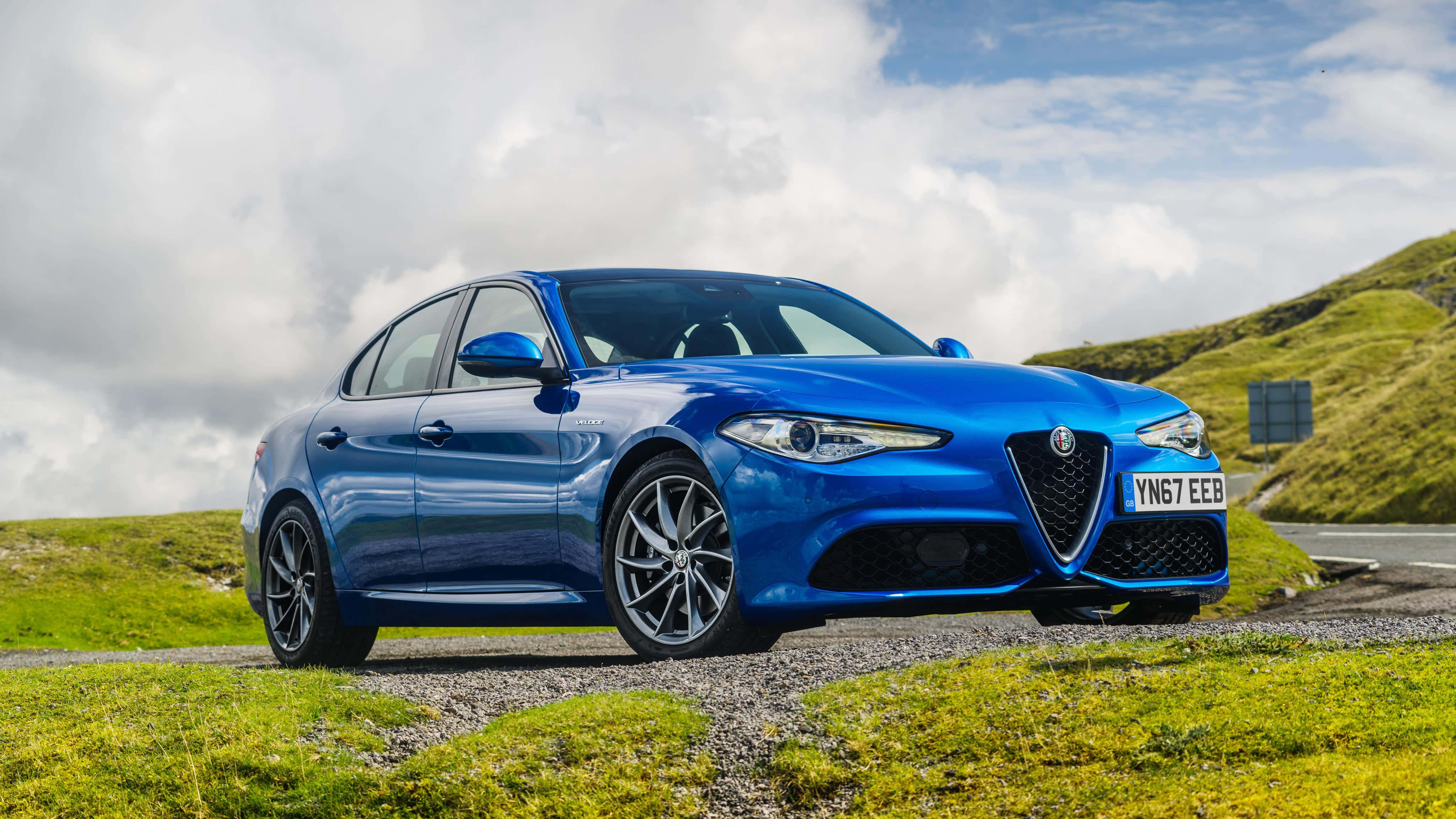 Alfa Romeo Giulia 桌布 壁紙 — 一架充滿活力的藍色愛快羅密歐房車停泊在碎石路上，旁邊是草地丘陵，天空多雲。