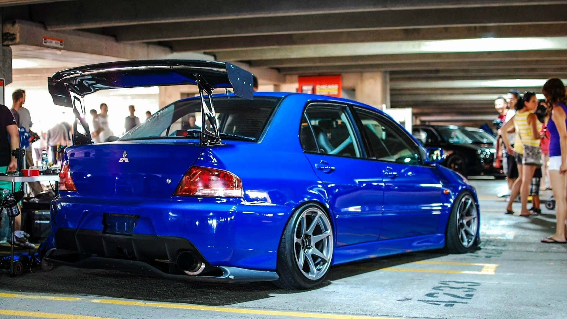JDM 車 桌布 壁紙 — 一架充滿活力的藍色改裝三菱 Lancer Evolution，帶有大型後擾流板，停在混凝土停車場，附近有人。