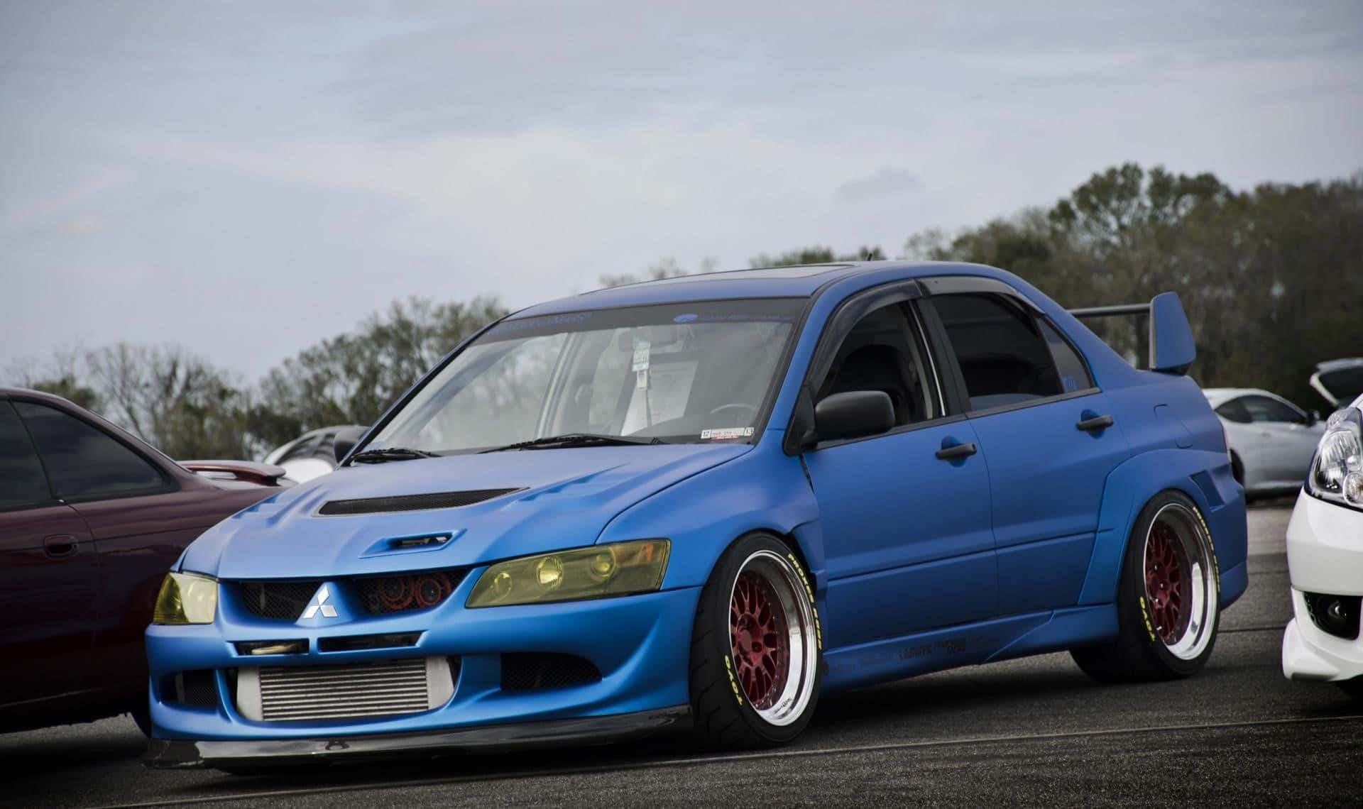 JDM 車 桌布 壁紙 — 一架充滿活力的藍色改裝三菱Lancer Evolution，配紅色輪圈，停泊在戶外停車場，天空多雲。