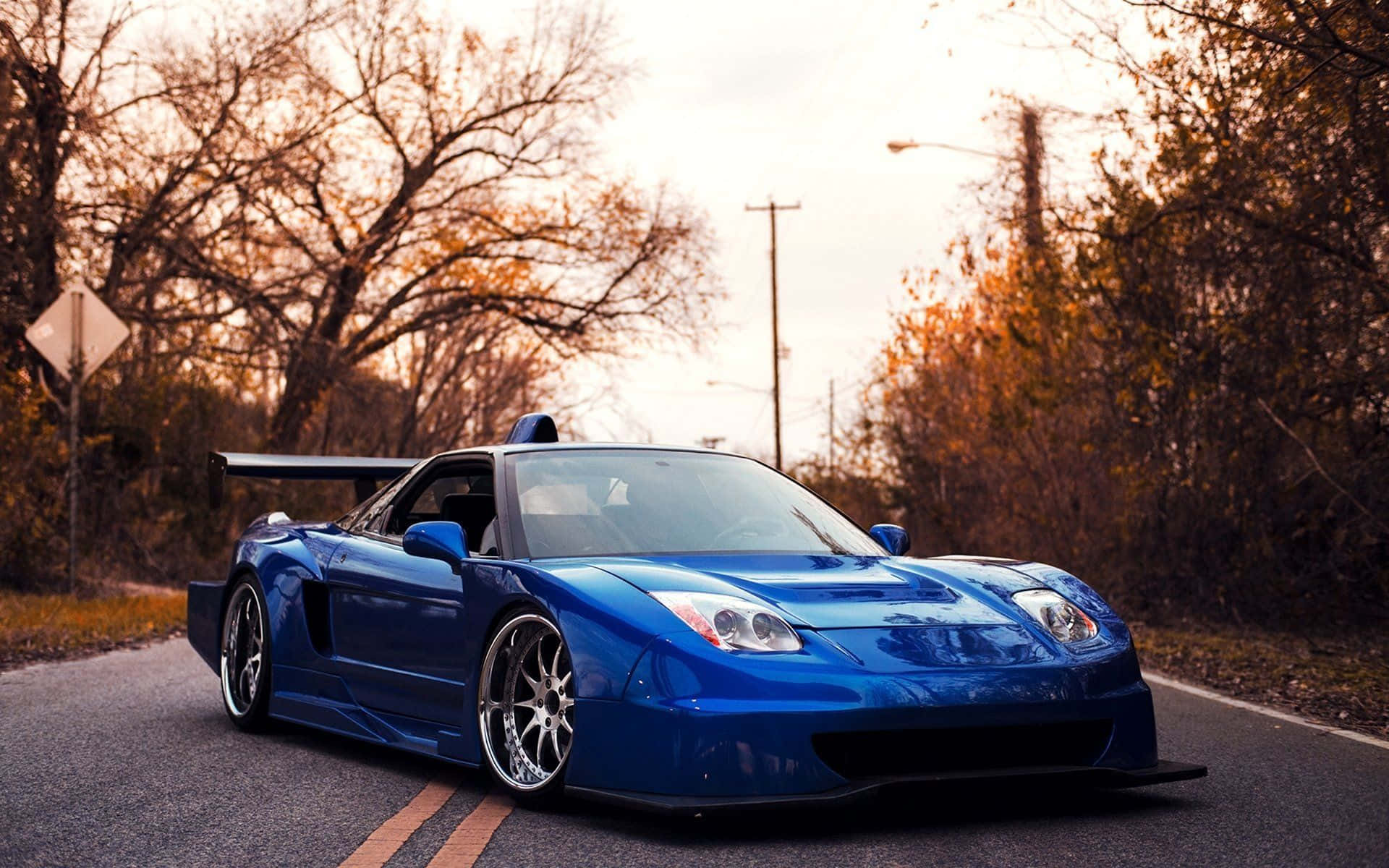 JDM 車 桌布 壁紙 — 一架充滿活力的藍色改裝跑車，帶有大型後擾流板，在黃金時刻停泊在鄉村道路上，周圍環繞著秋天的樹木。
