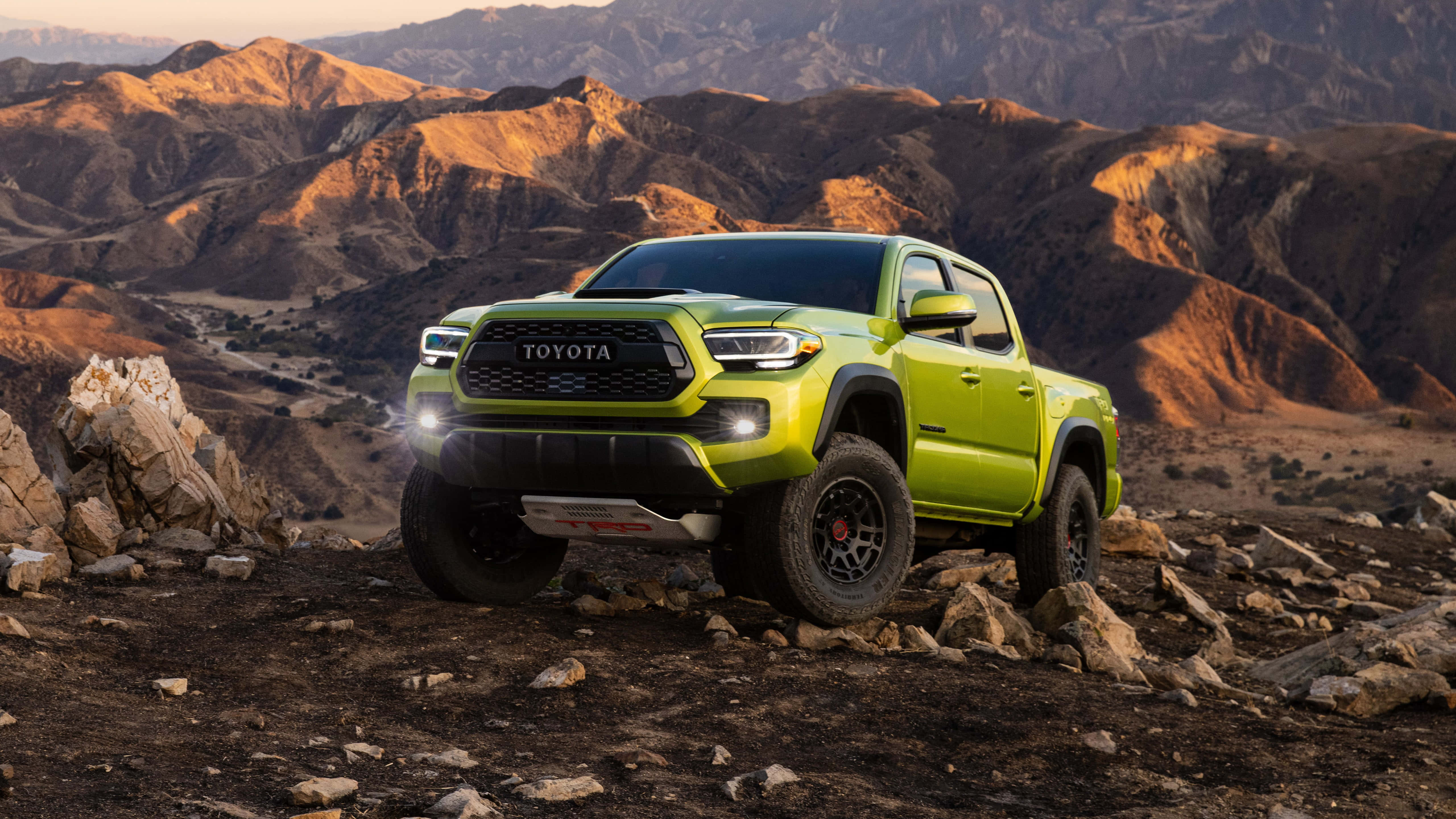Toyota 桌布 壁紙 — 一架充滿活力的青檸綠色豐田 Tacoma 皮卡車，在陽光照耀的沙漠山脈背景下，矗立在岩石地形上。