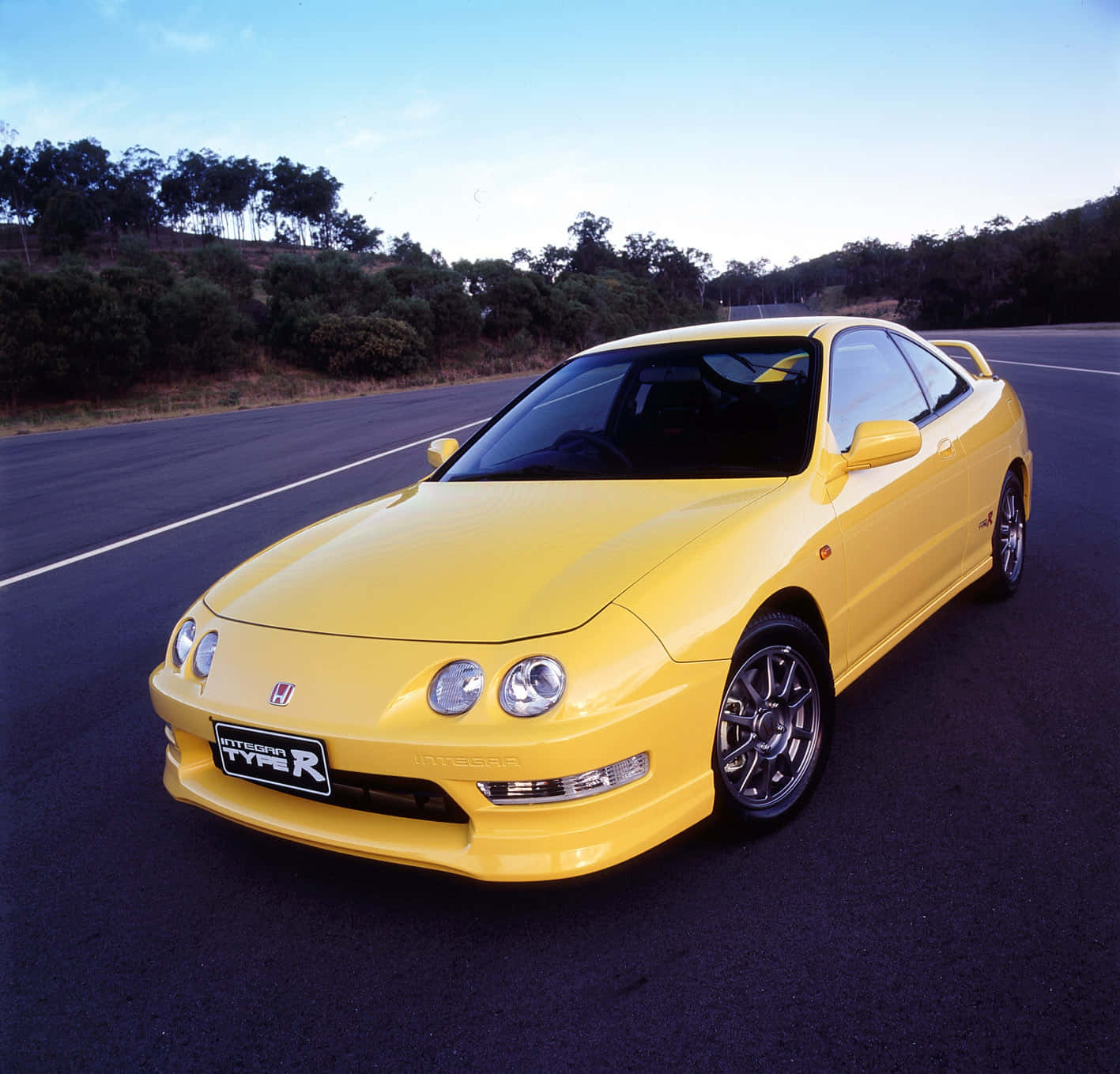 Honda Integra 桌布 壁紙 — 一架充滿活力的黃色本田Integra Type R跑車停泊在空曠的瀝青路上，背景有樹木和山丘，天空清澈湛藍。