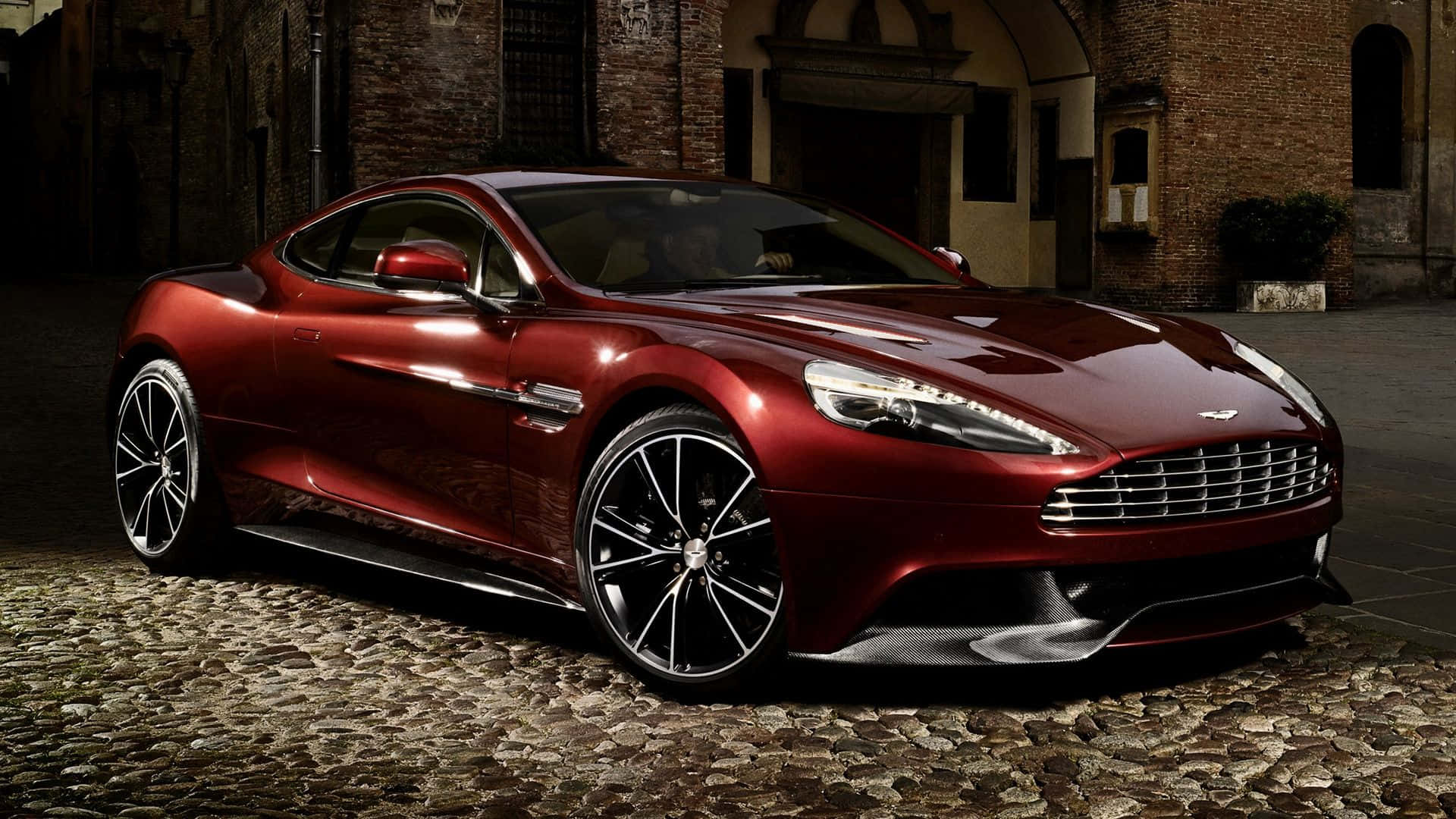 Aston Martin Vanquish 桌布 壁紙 — 一架光亮的紅色Aston Martin跑車在戲劇性燈光下，在歷史磚砌建築旁的鵝卵石街道上閃閃發光。