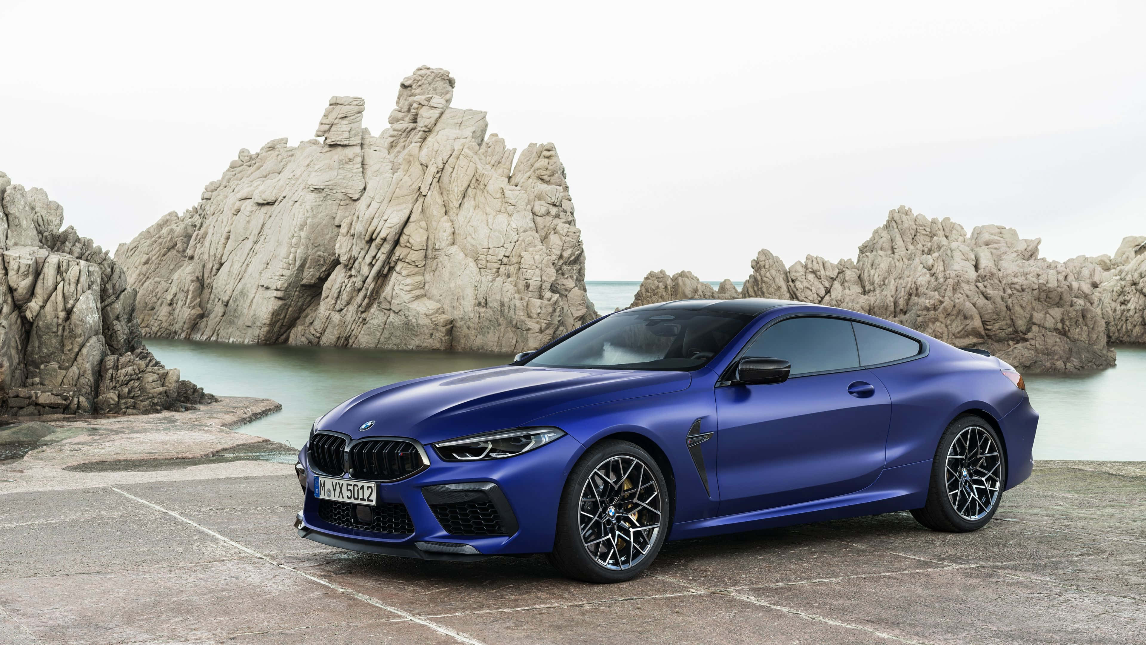 4K BMW 壁紙 桌布 壁紙 — 一架光亮的藍色BMW M8 Coupe停泊在石碼頭上，旁邊是崎嶇的海岸懸崖和風平浪靜的大海，天空多雲。