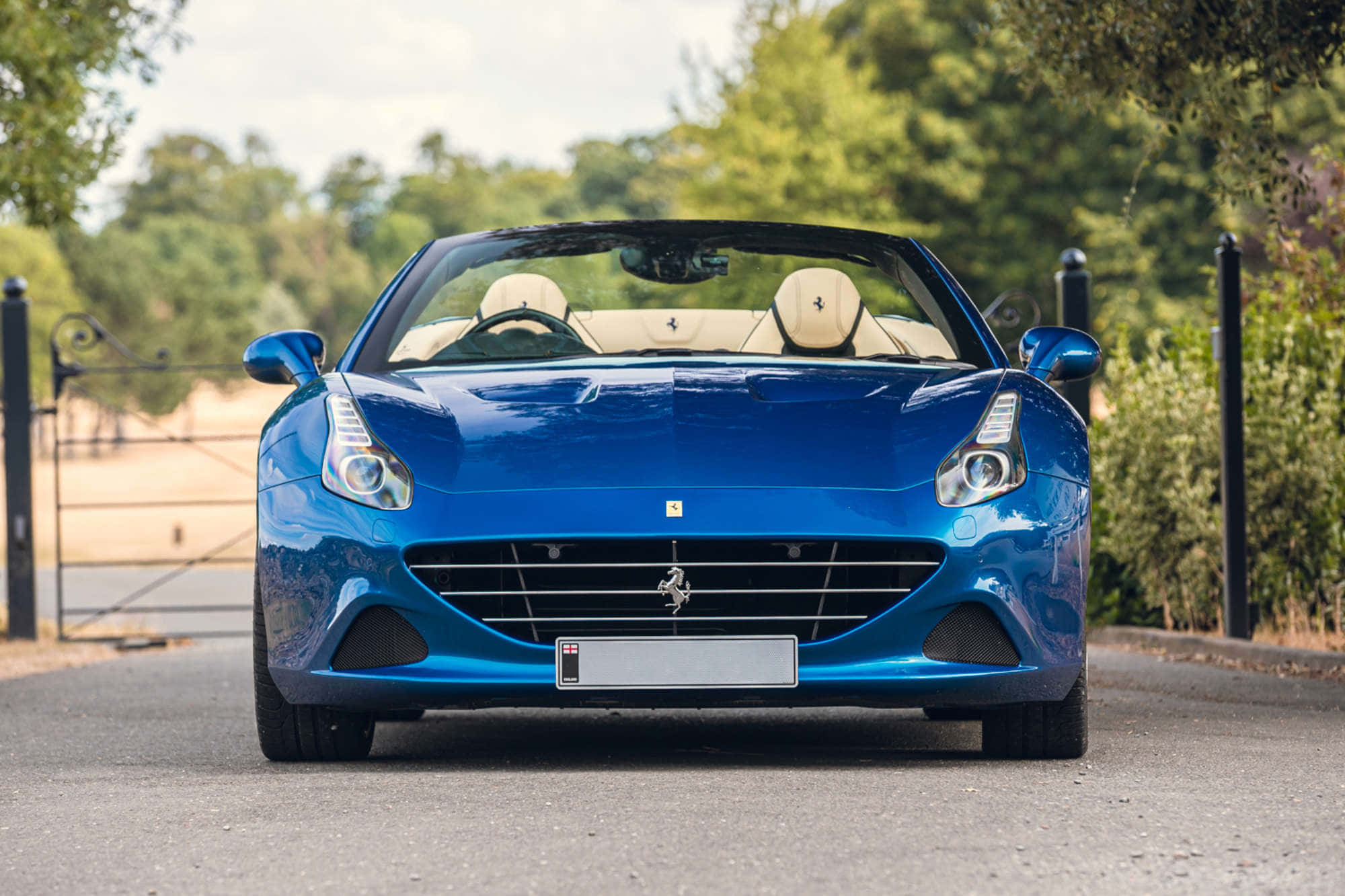 Ferrari California T 桌布 壁紙 — 一架光亮的藍色法拉利開篷車停泊在鋪砌的道路上，背景有茂密的綠樹和一個鍛鐵大門。