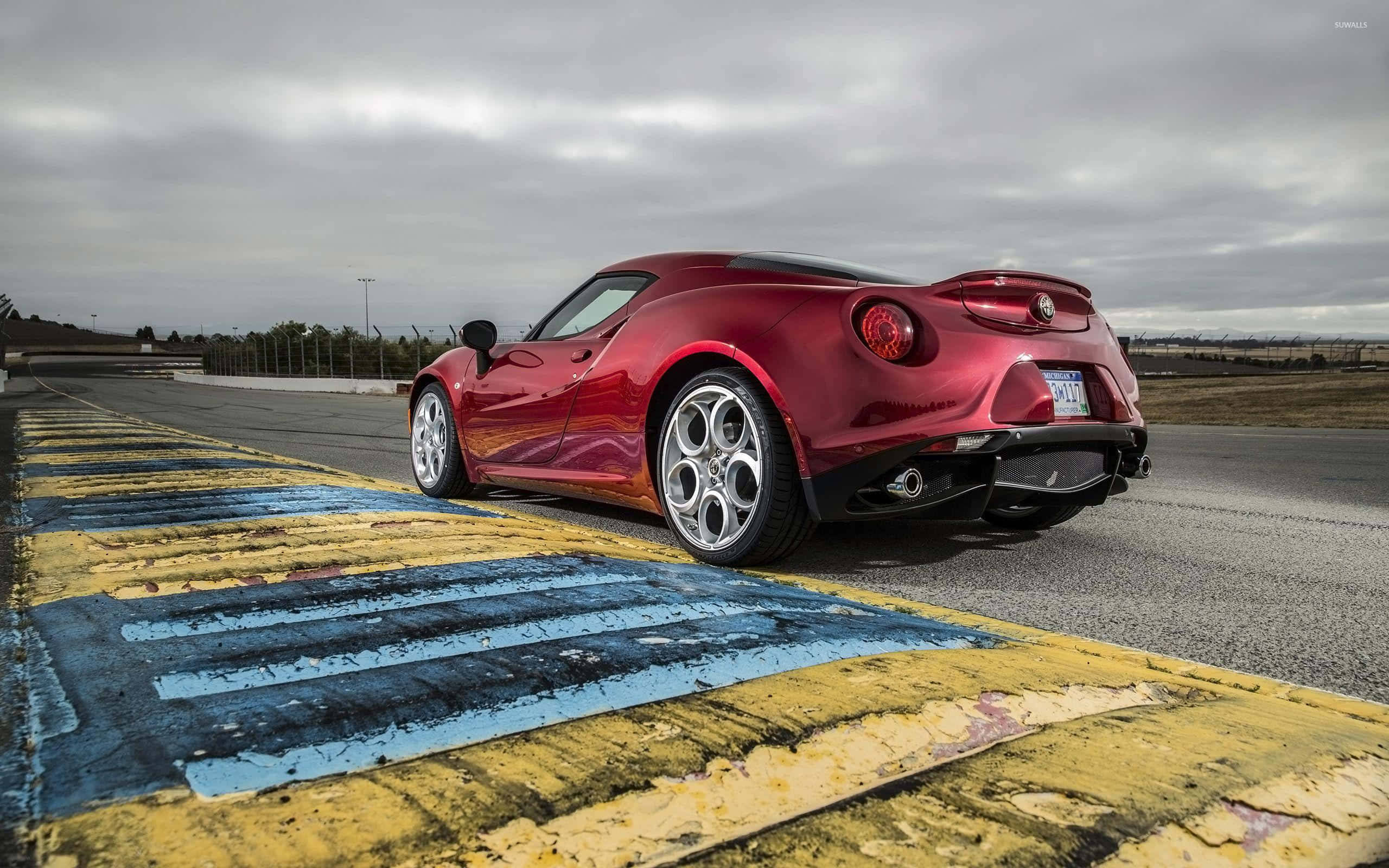 Alfa Romeo 4c 桌布 壁紙 — 一架光澤的紅色Alfa Romeo 4C跑車停泊在風化的黃藍色賽道邊緣，天空多雲。