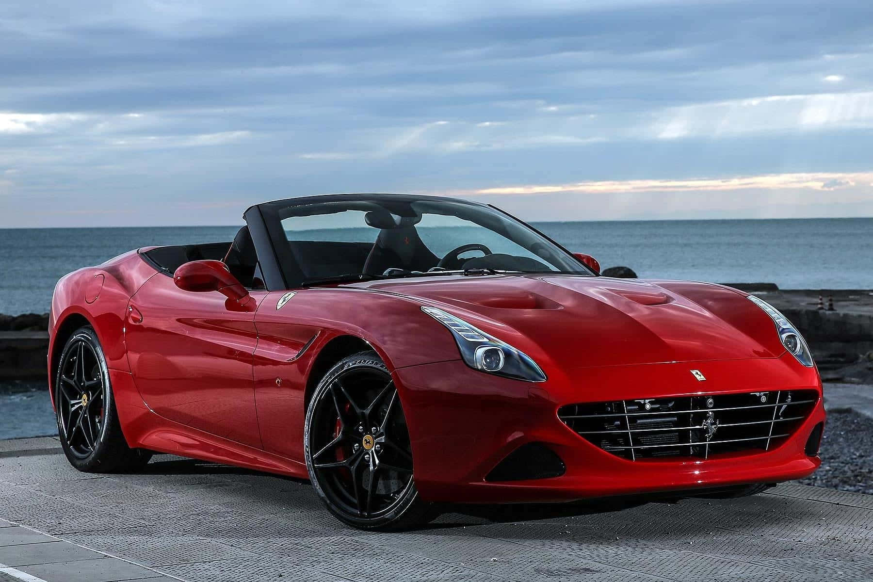 Ferrari California T 桌布 壁紙 — 一架光澤的紅色法拉利開篷車停泊在石碼頭上，背景是海洋和多雲的天空。
