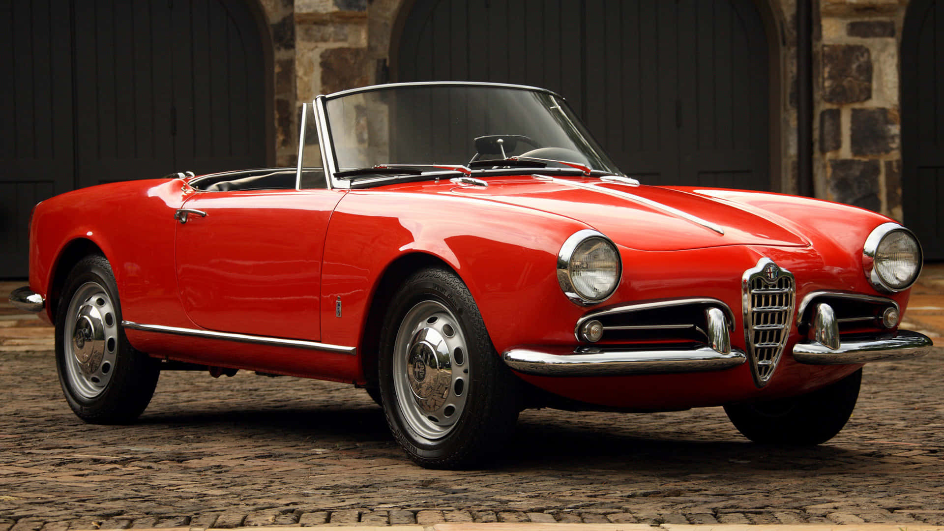 Alfa Romeo Spider 桌布 壁紙 — 一架光澤的紅色經典愛快羅密歐Spider開篷車停在鵝卵石街道上，背景有石拱門。