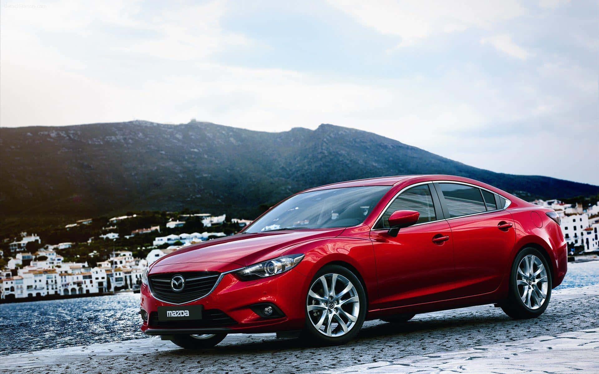 Mazda 6 桌布 壁紙 — 一架光澤的紅色萬事得房車停泊在鵝卵石海濱，背景有白色建築物和山脈。