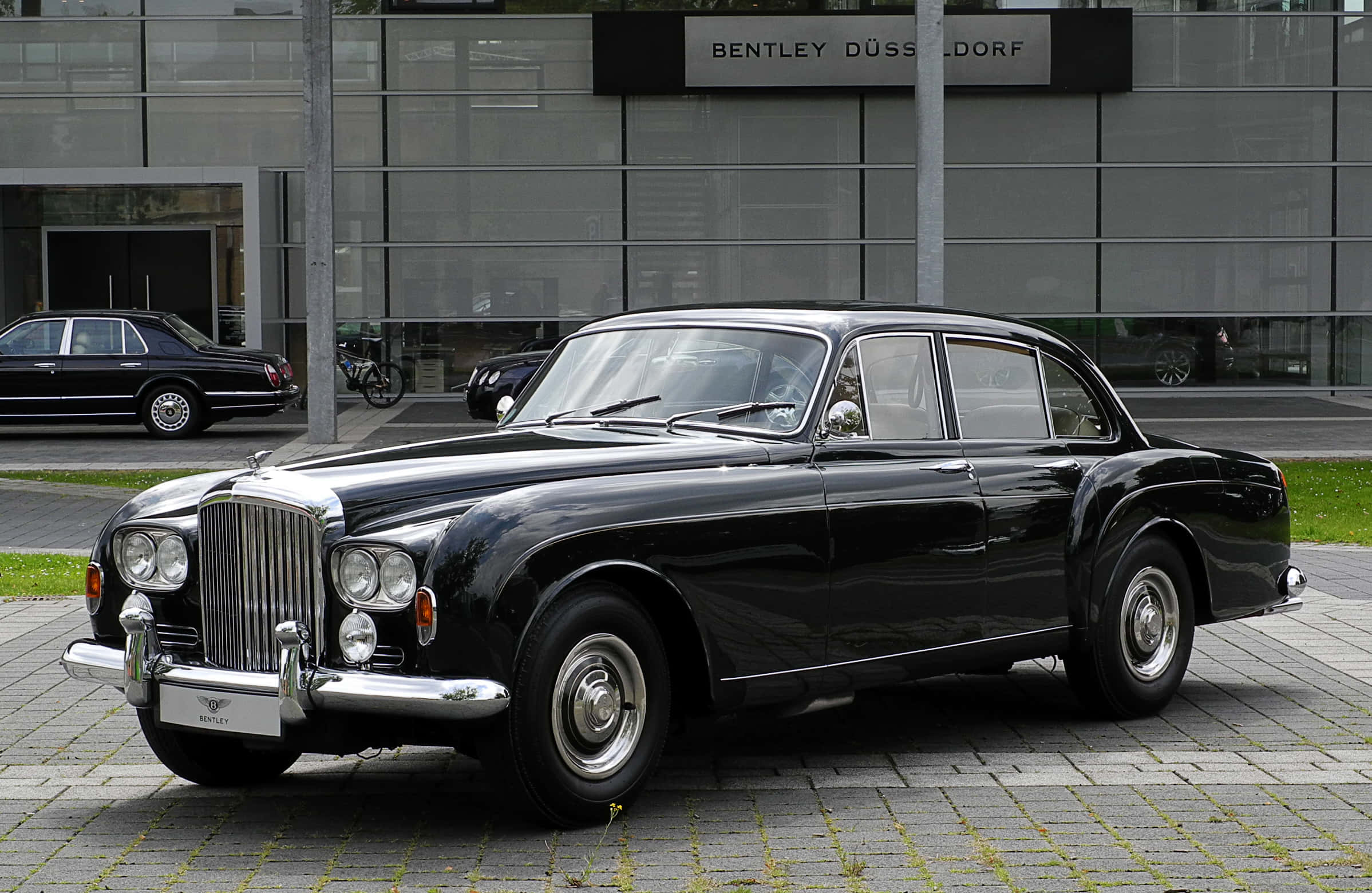 Bentley S3 桌布 壁紙 — 一架光澤黑色復古賓利房車停泊在現代玻璃建築前的鋪砌庭院。