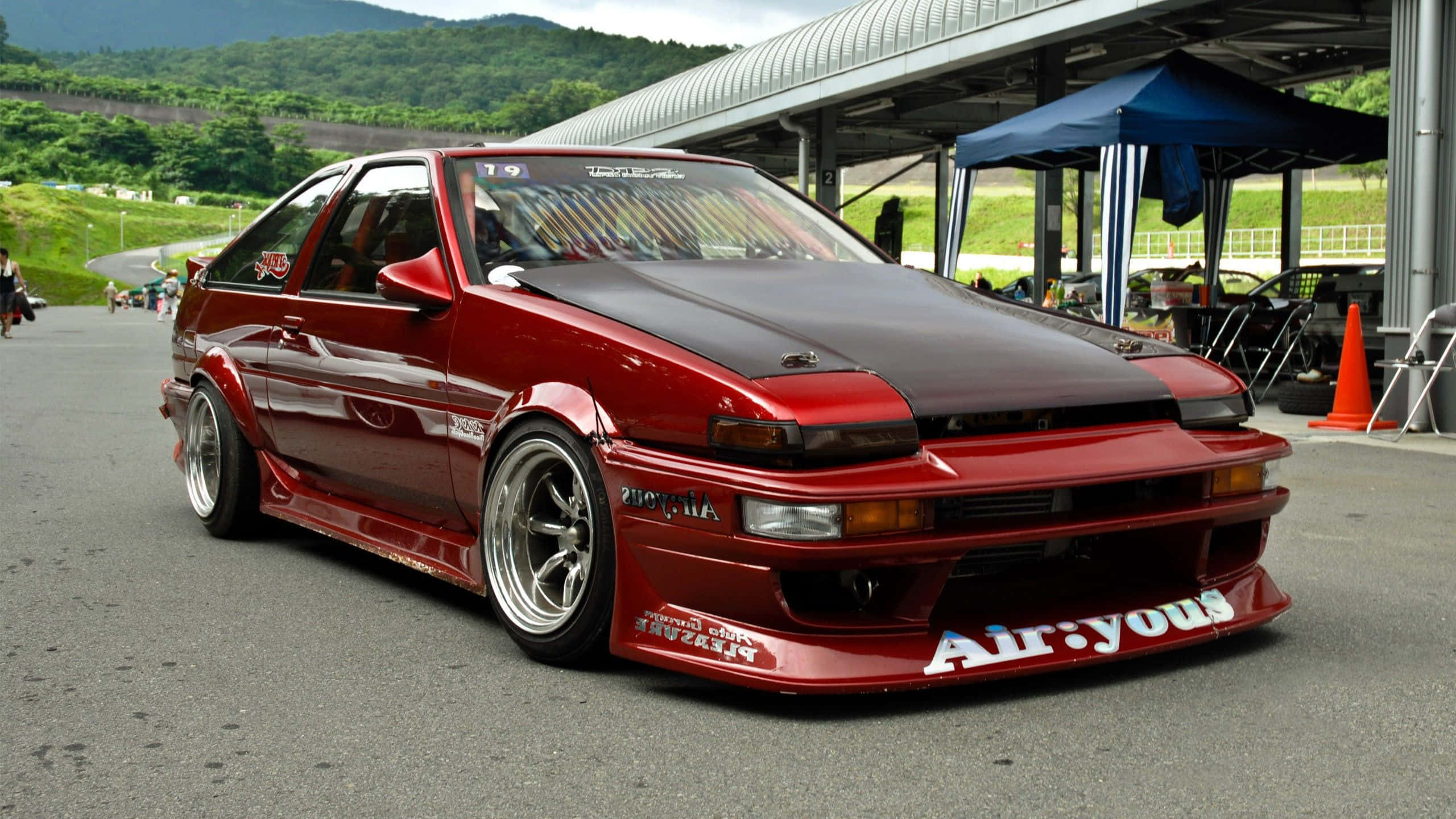 Jdm Car 桌布 壁紙 — 一架光面紅色改裝AE86跑車，配備自訂寬體套件及「Air:yous」貼紙，停泊在賽車場維修區。