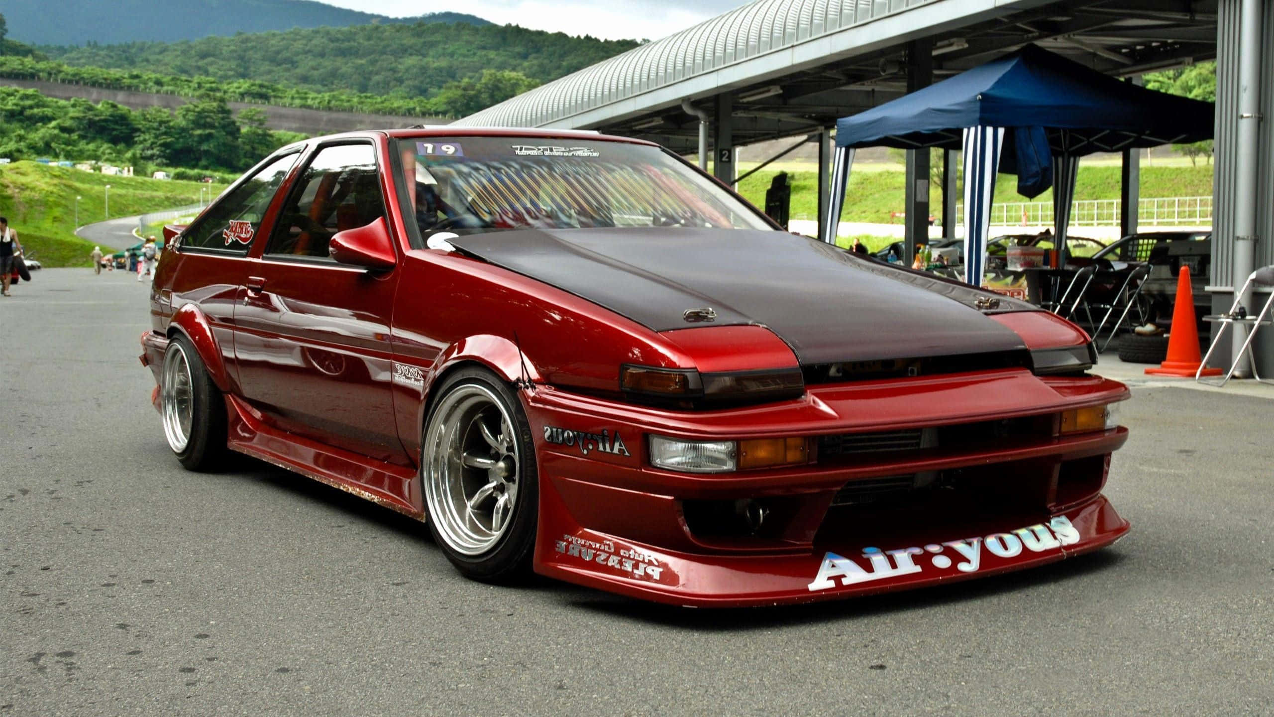 JDM 桌布 壁紙 — 一架光面紅色改裝AE86跑車，配備寬體車身套件和訂製輪圈，停泊在賽車場維修區。