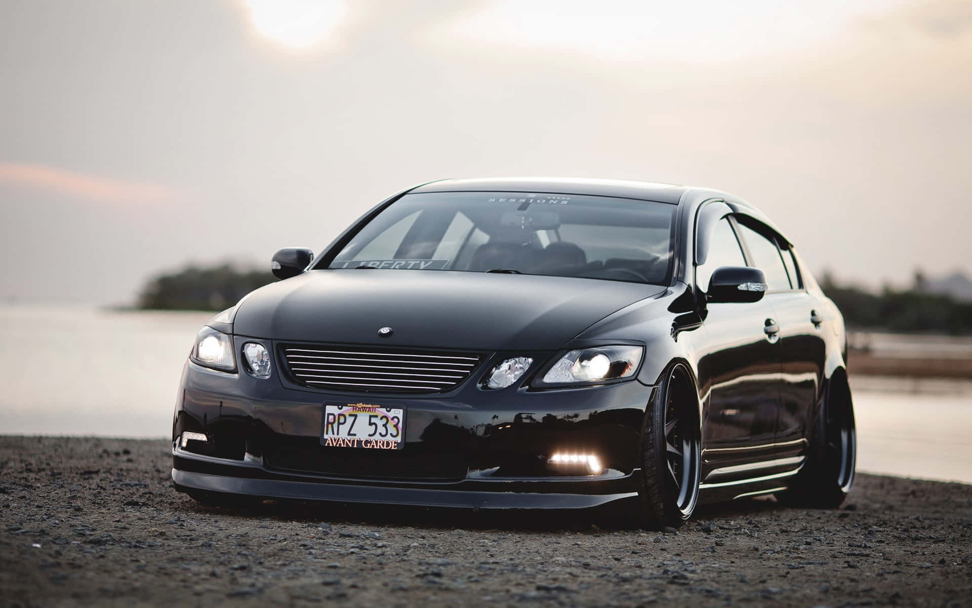 Lexus Gs 桌布 壁紙 — 一架光面黑色改裝房車，帶有發光車頭燈，停在日落時的礫石岸邊。