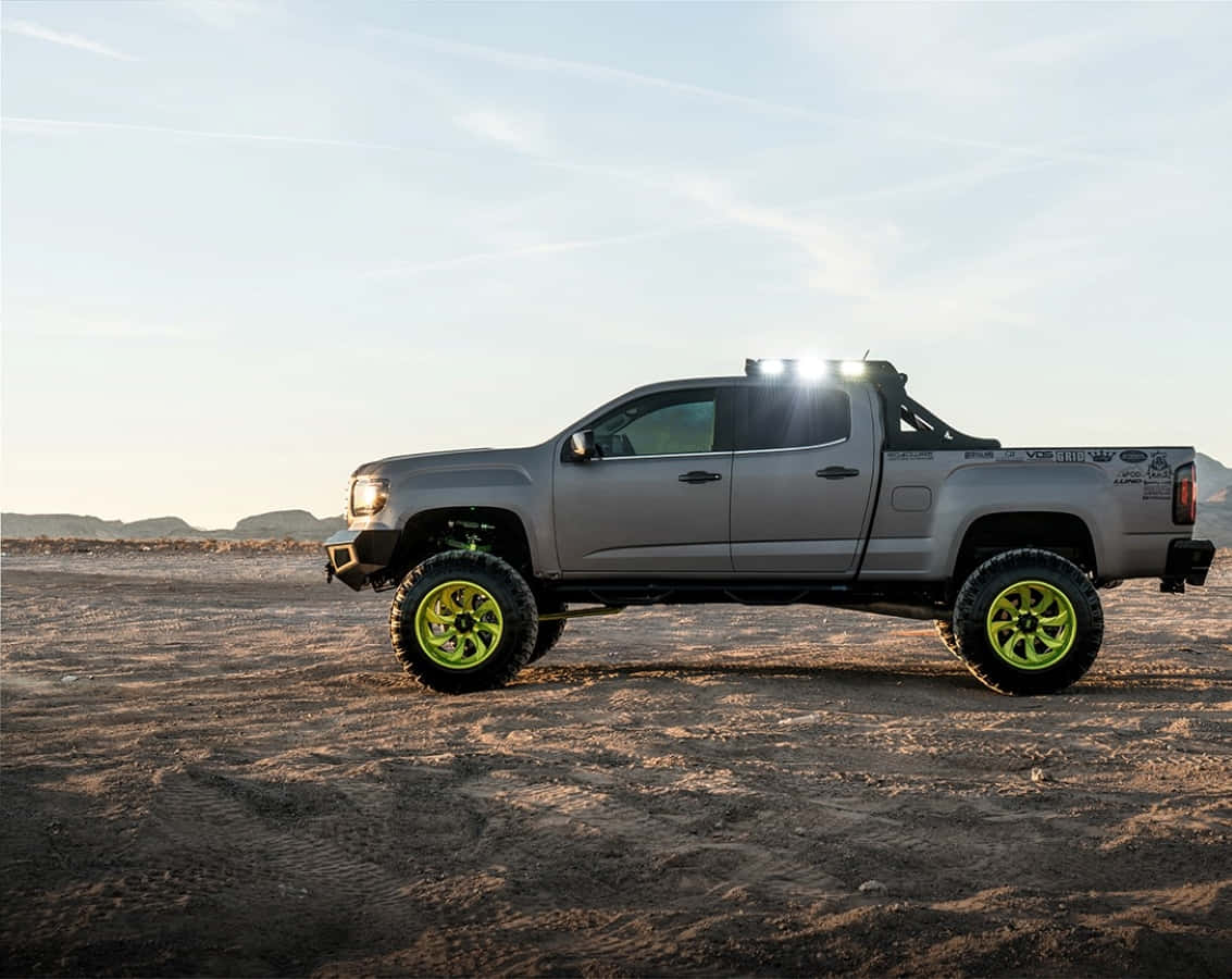 Gmc Canyon 桌布 壁紙 — 一架升高嘅灰色皮卡車配螢光綠色車輪，喺晴朗天空下企喺陽光普照嘅沙漠地形上。