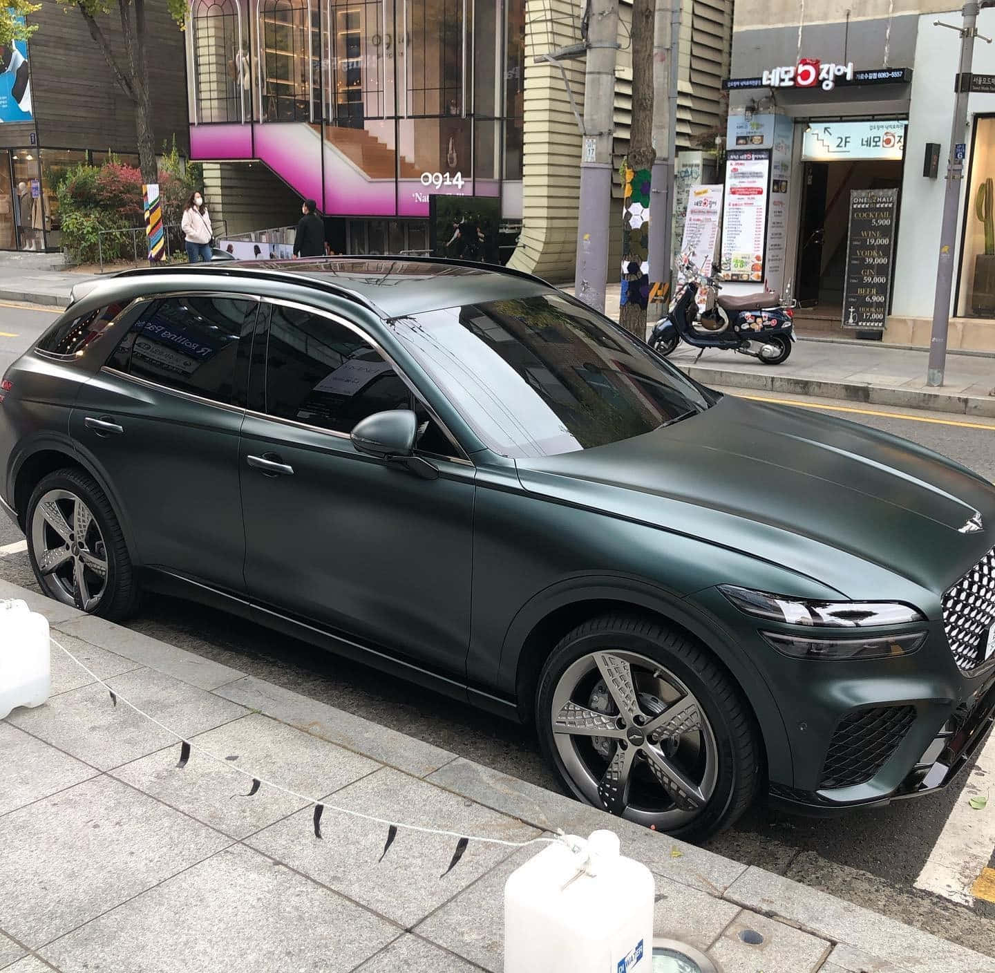 Genesis Gv70 桌布 壁紙 — 一架啞光深綠色SUV停泊喺城市行人路旁邊，隔籬有現代建築同埋一架滑板車。