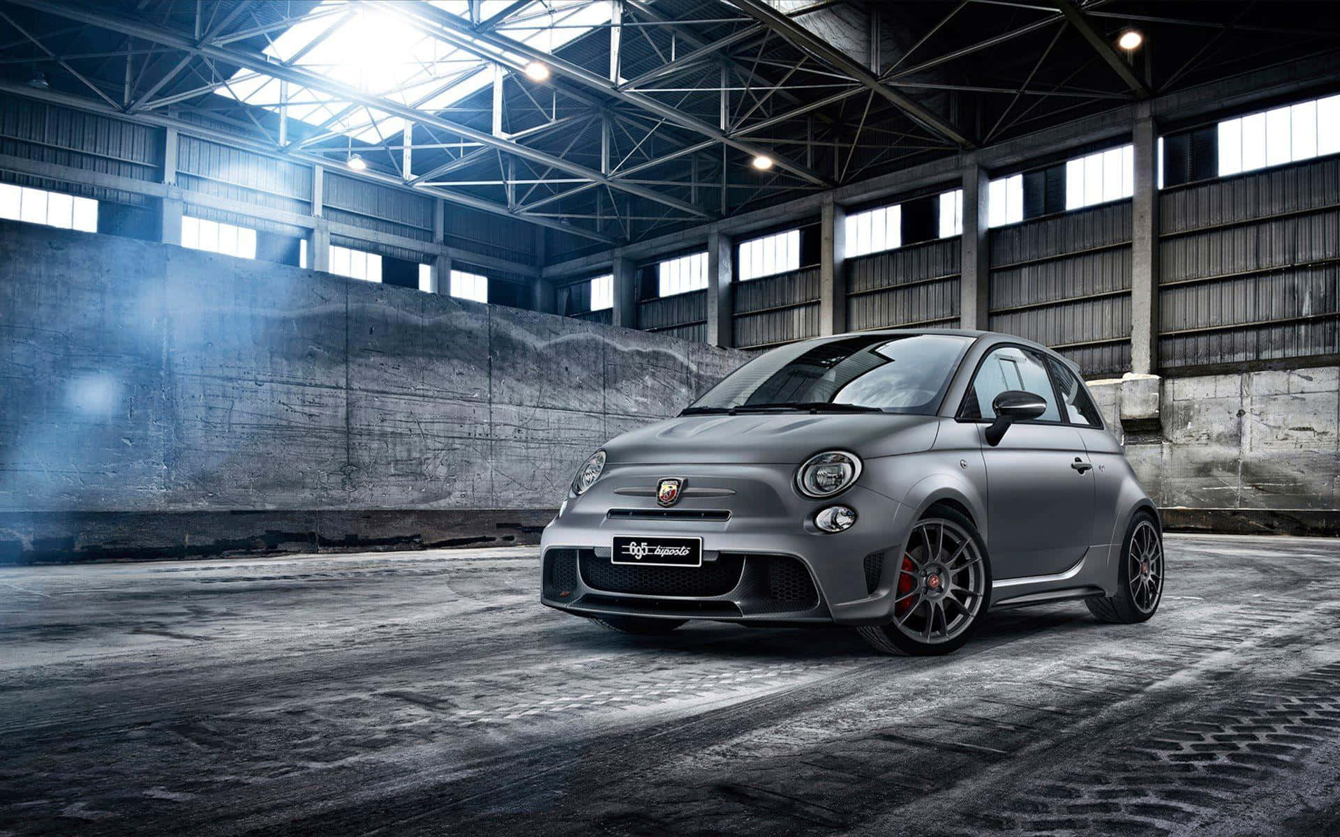 Abarth 桌布 壁紙 — 一架啞光灰色 Fiat 500 Abarth 跑車配紅色制動卡鉗，戲劇性地停放在一個充滿灰塵、高聳鋼樑和光線照射的工業倉庫中。