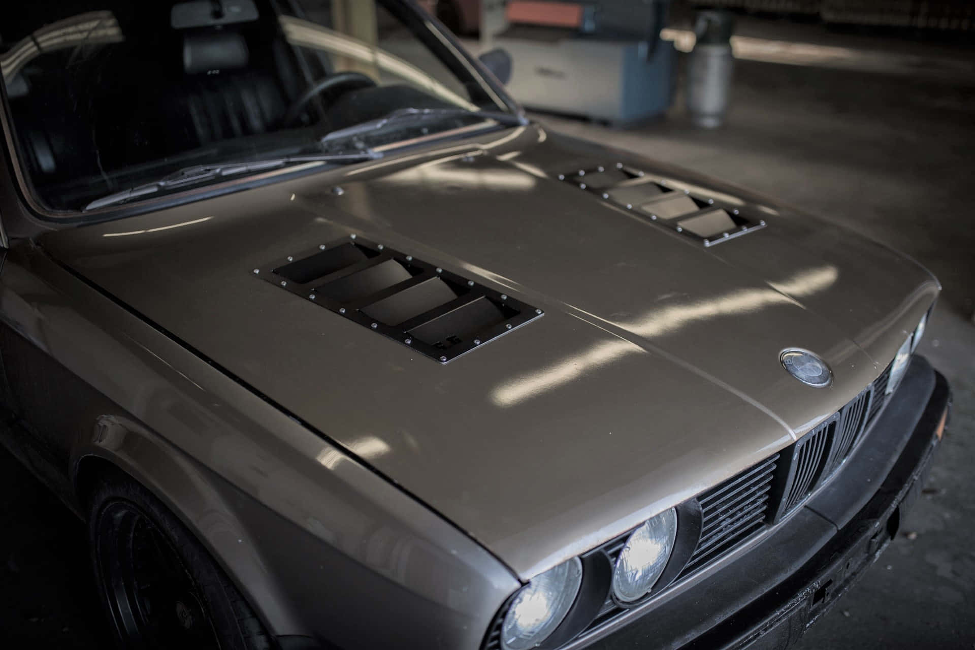 Car Hood 桌布 壁紙 — 一架啞光灰色BMW E30，帶黑色引擎蓋進氣口，停放在昏暗的車庫中，沐浴在自然光線中。