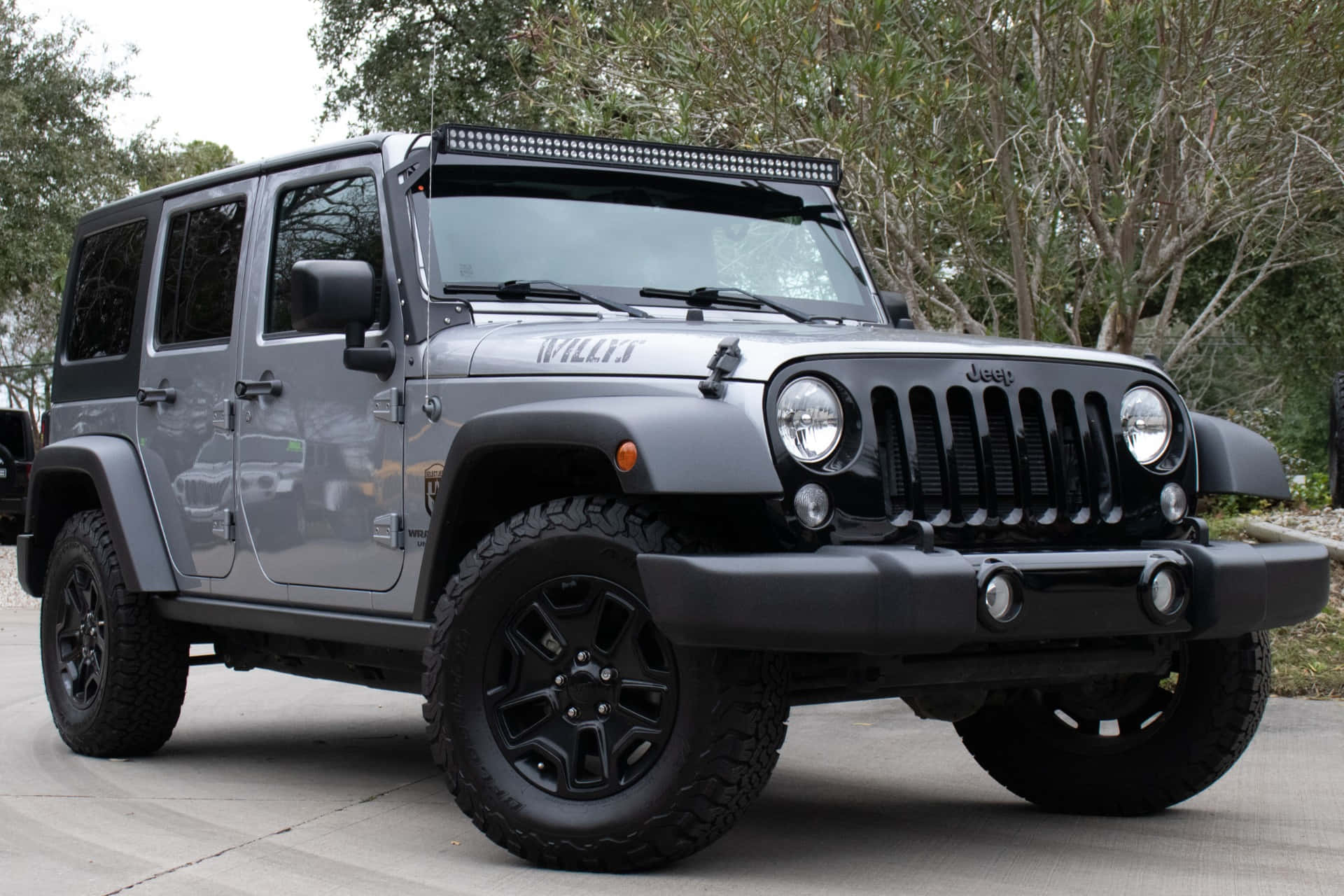 Jeep Willys 桌布 壁紙 — 一架啞光灰色Jeep Wrangler，帶有黑色飾件和越野輪胎，停泊在樹木環繞的混凝土車道上。