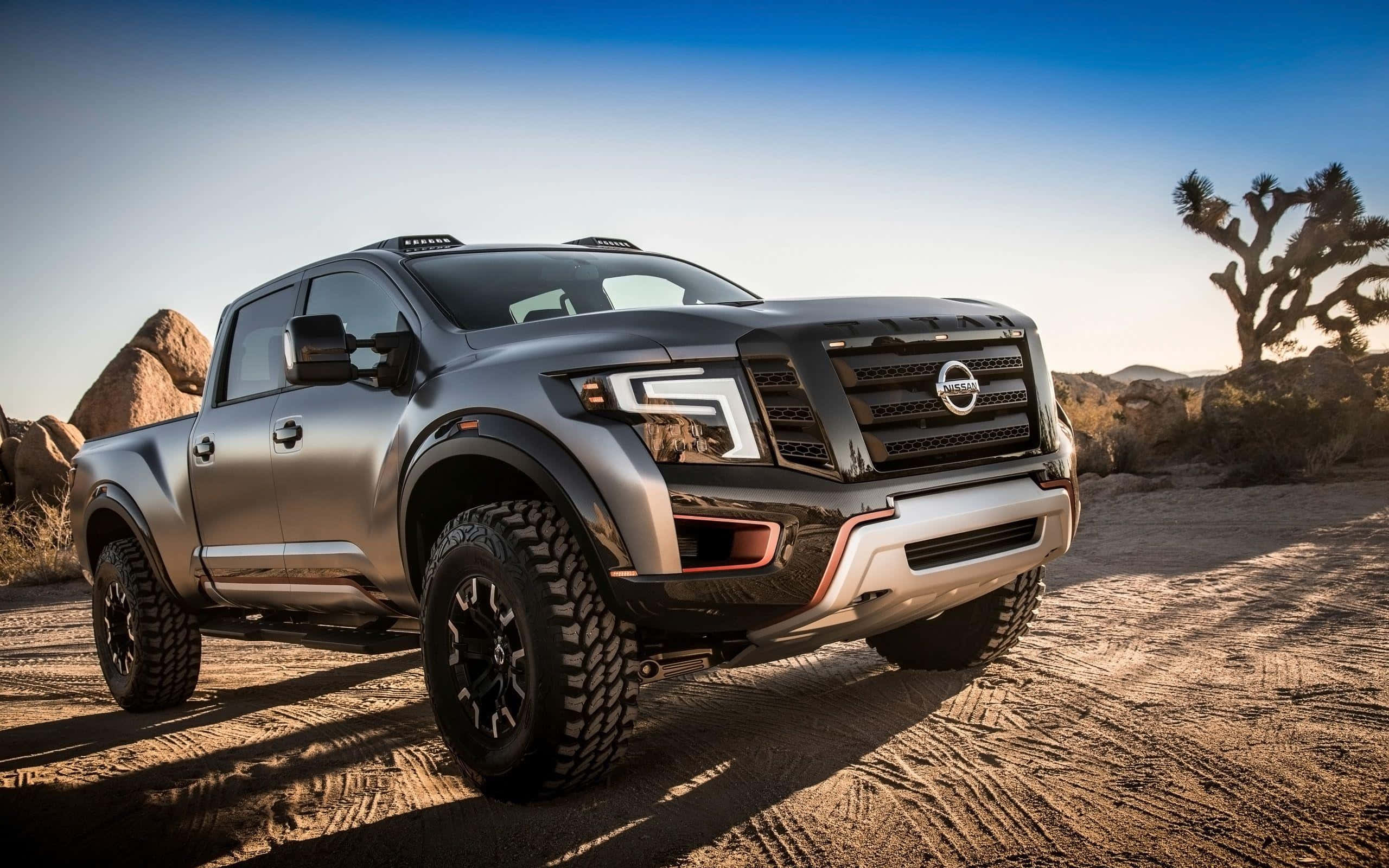 Nissan Titan 桌布 壁紙 — 一架啞光灰色日產Frontier貨車，帶有黑色飾邊和大輪胎，停在沙質沙漠地形上，背景是晴朗的藍天和一棵約書亞樹。