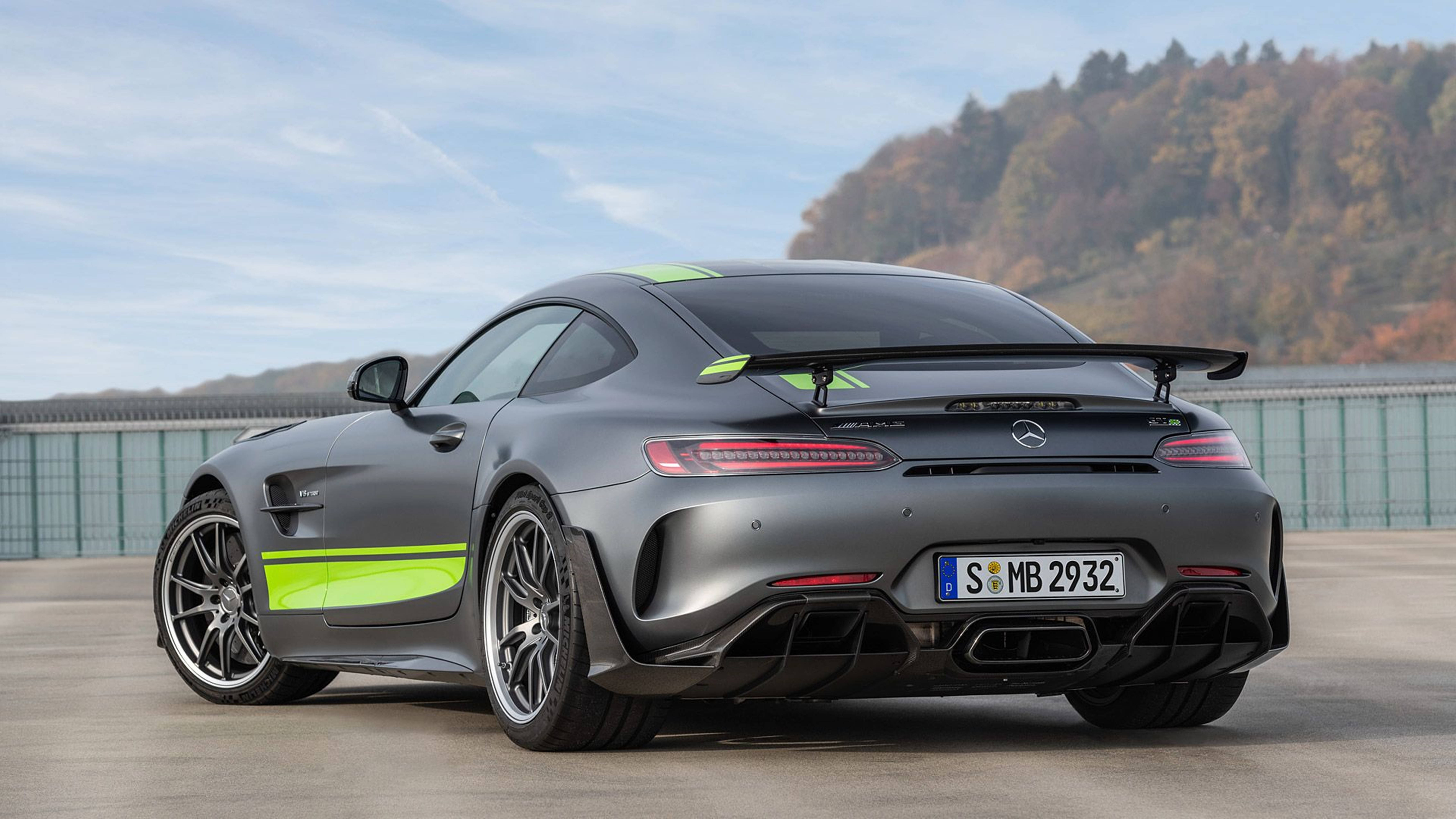 Sports Car 桌布 壁紙 — 一架啞光灰色梅賽德斯-AMG GT R 跑車，帶有螢光綠色點綴，停泊在混凝土場地上，背景是樹木繁茂的山丘，天空清澈湛藍。