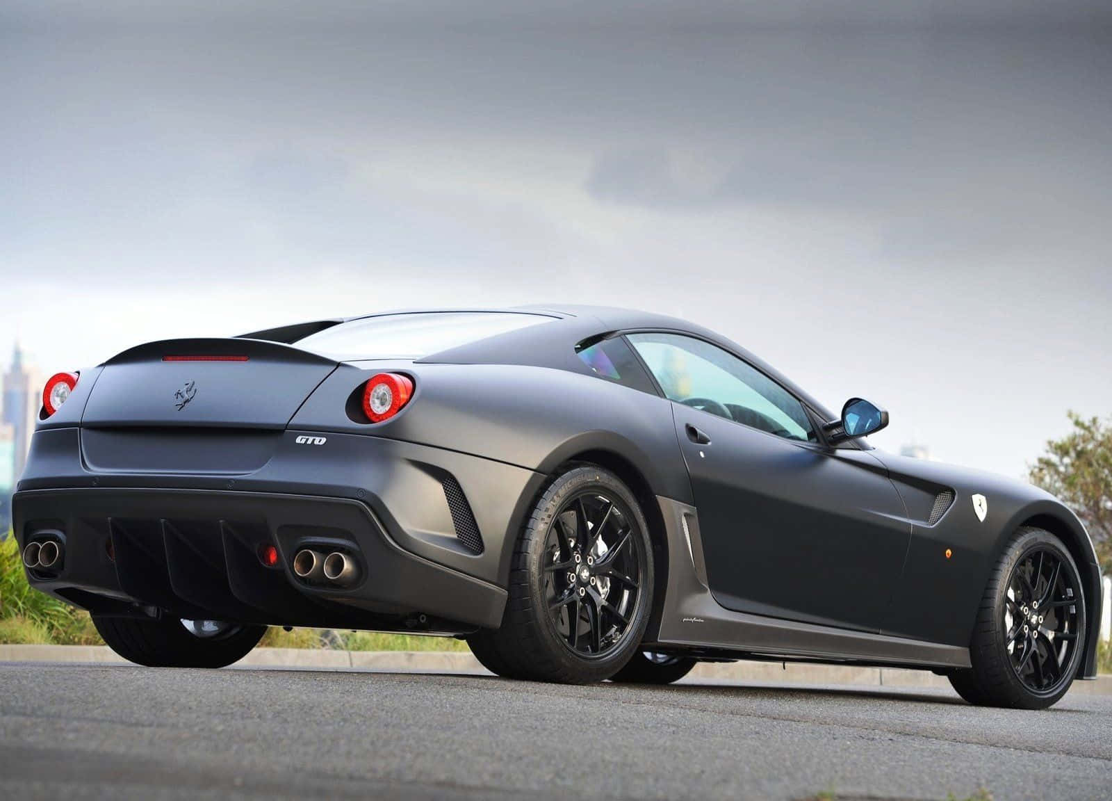 Ferrari 599 Gtb Fiorano 桌布 壁紙 — 一架啞光灰色法拉利 599 GTO 跑車停泊在瀝青路上，天空多雲，遠處有城市景觀。