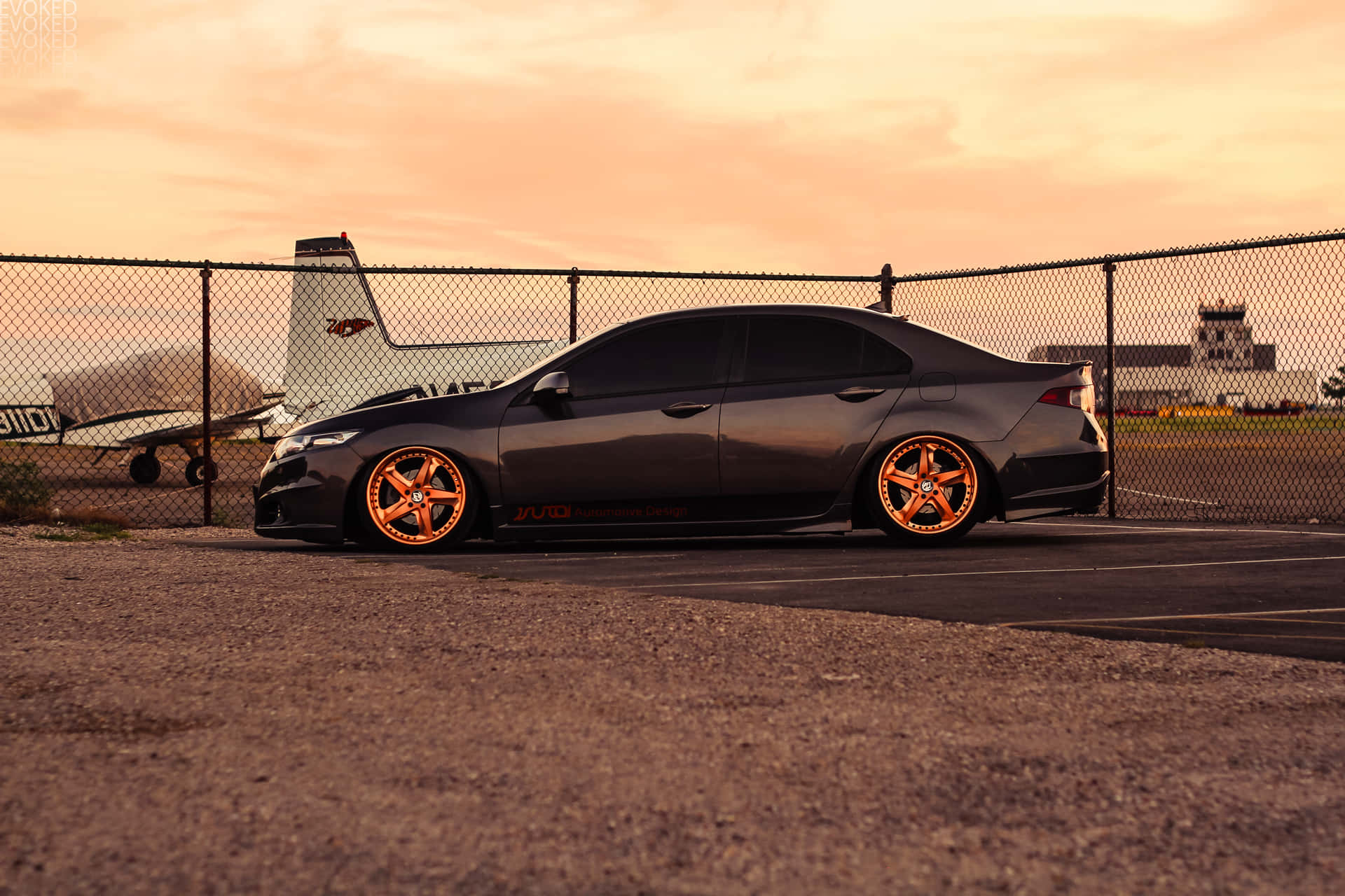 Acura Tsx 桌布 壁紙 — 一架啞光灰色轎車，配備發光橙色輪圈，停泊在日落時分的碎石停車場，背景有鐵絲網圍欄和飛機。