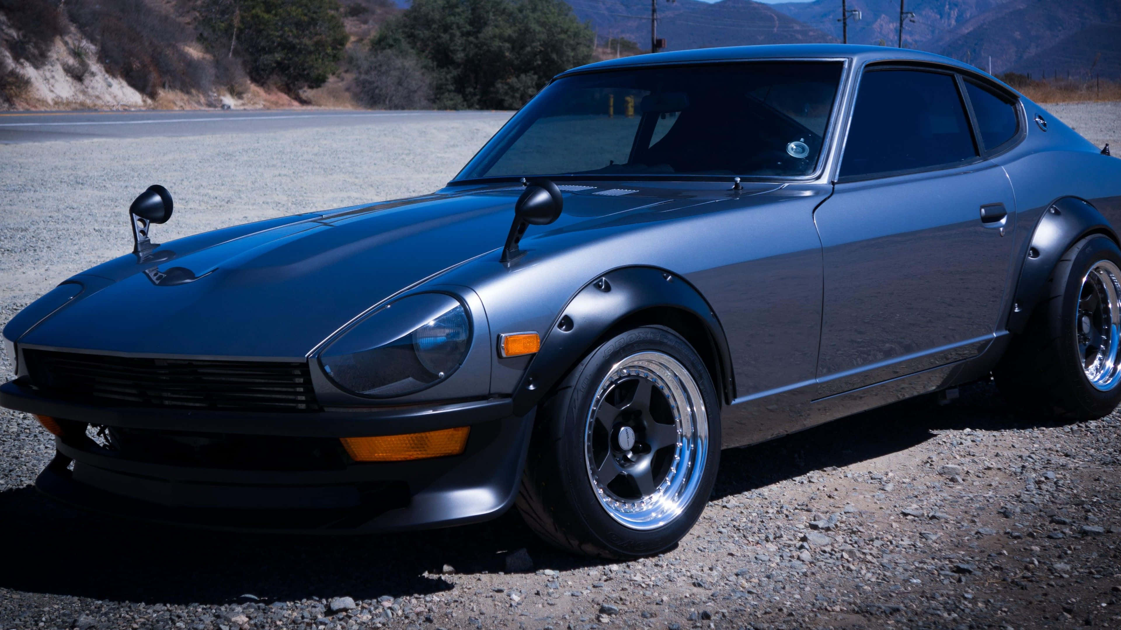 Datsun 桌布 壁紙 — 一架啞光藍色經典跑車配備定制輪圈，在明亮日光下停泊在沙漠道路旁的礫石上。