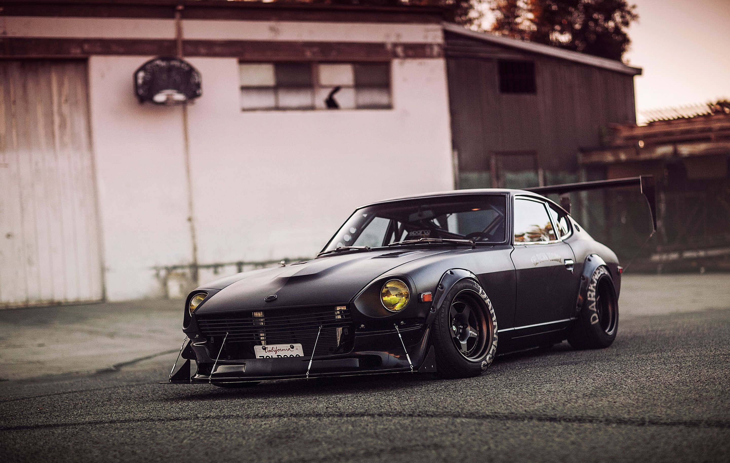 Datsun 桌布 壁紙 — 一架啞光黑色改裝Datsun 240Z，配備寬輪胎和後尾翼，在黃昏時停放在風化車庫前。
