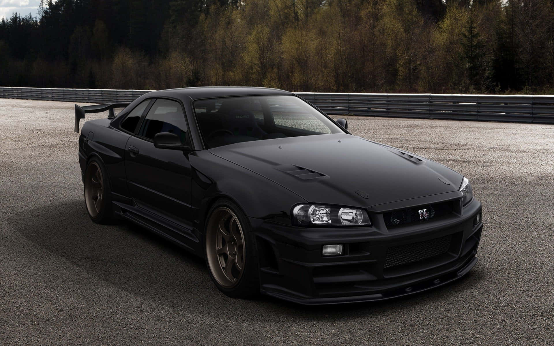 啞光黑色 桌布 壁紙 — 一架啞光黑色日產Skyline GT-R停泊在賽道上，背景是樹木繁茂的山丘。