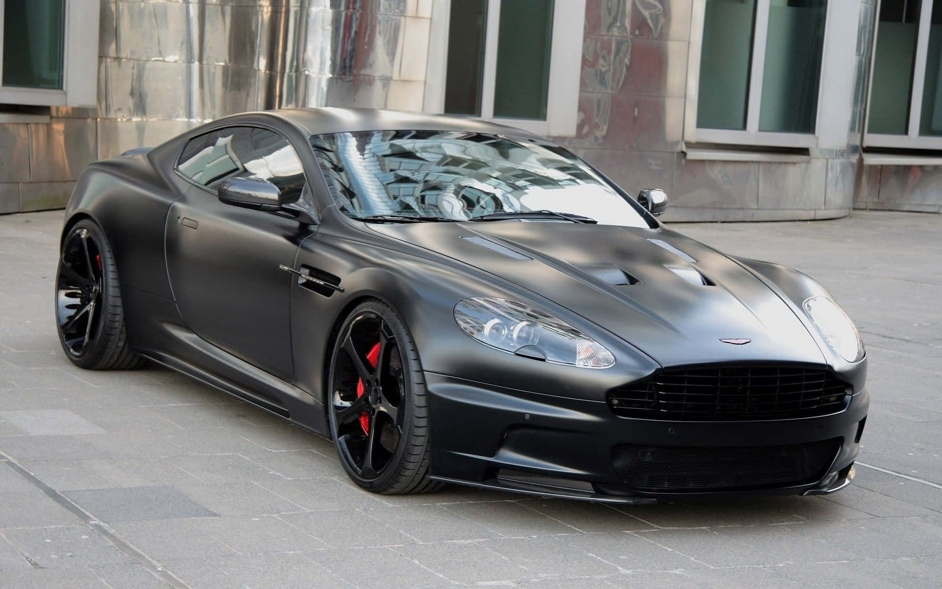 Aston Martin Db9 桌布 壁紙 — 一架啞黑色 Aston Martin 跑車配紅色制動卡鉗，停泊在瓷磚鋪設的城市行人路上。