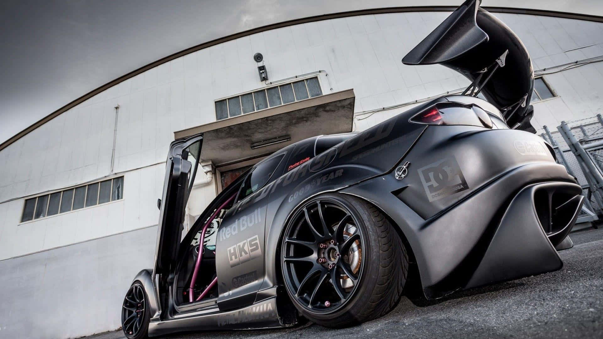 Mazda Rx-8 桌布 壁紙 — 一架啞黑色改裝跑車，車門打開，帶有大型尾翼，停泊在彎曲的工業建築前。