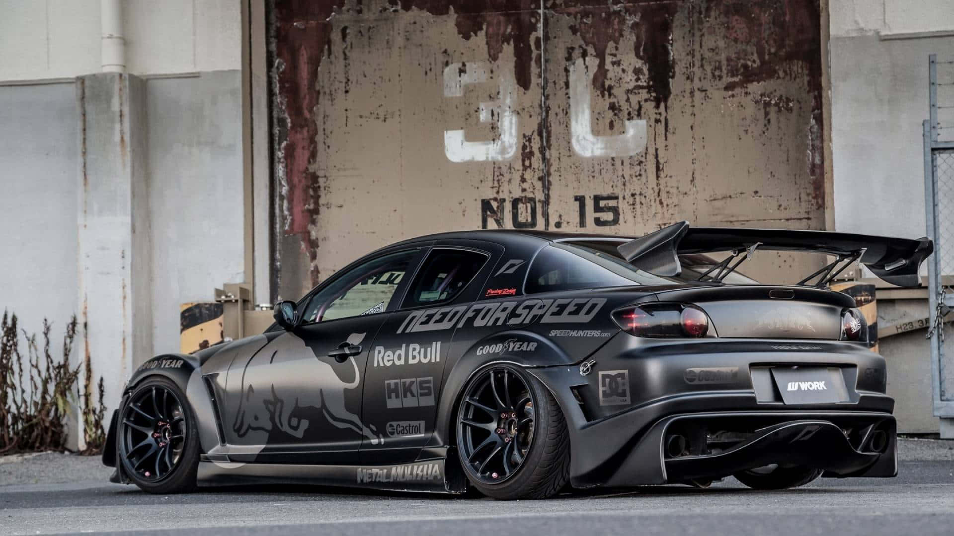 Mazda Rx-8 桌布 壁紙 — 一架啞黑色飄移車，帶有Red Bull和Need for Speed貼紙，停泊在風化的工業牆前。