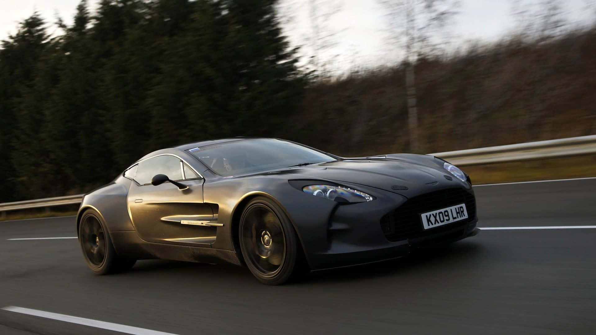 Aston Martin One-77 桌布 壁紙 — 一架型格啞黑色Aston Martin跑車在樹林環繞的道路上飛馳，背景呈現動態模糊效果。
