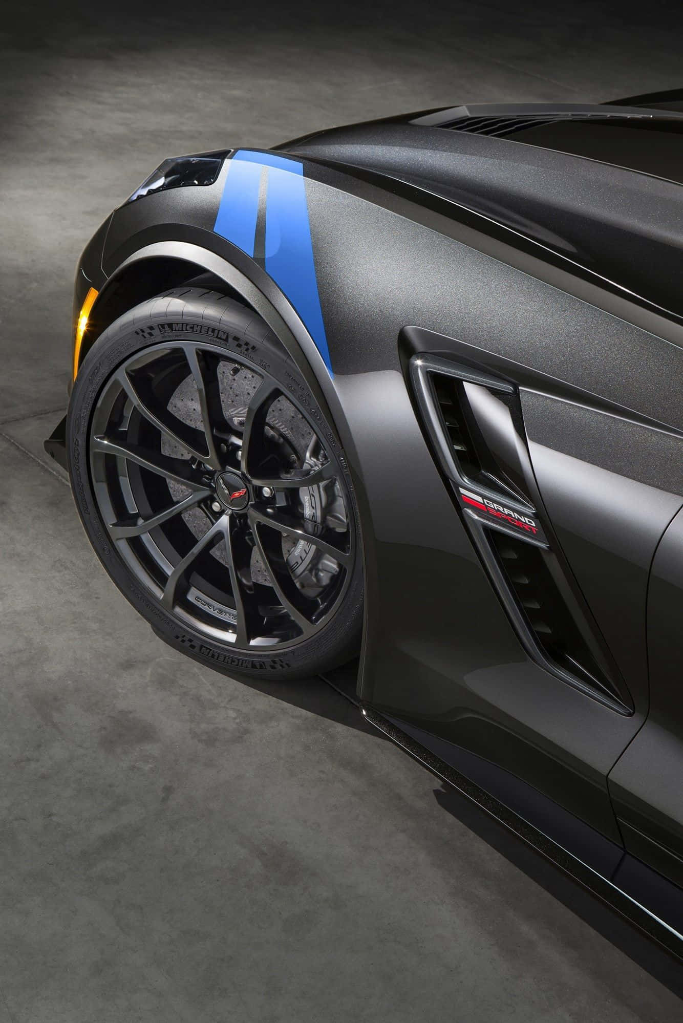 Chevrolet Corvette Grand Sport 桌布 壁紙 — 一架型格啞黑色跑車，配以大膽嘅藍色賽車條紋同精緻嘅多輻輪圈，喺昏暗嘅車庫中展示。