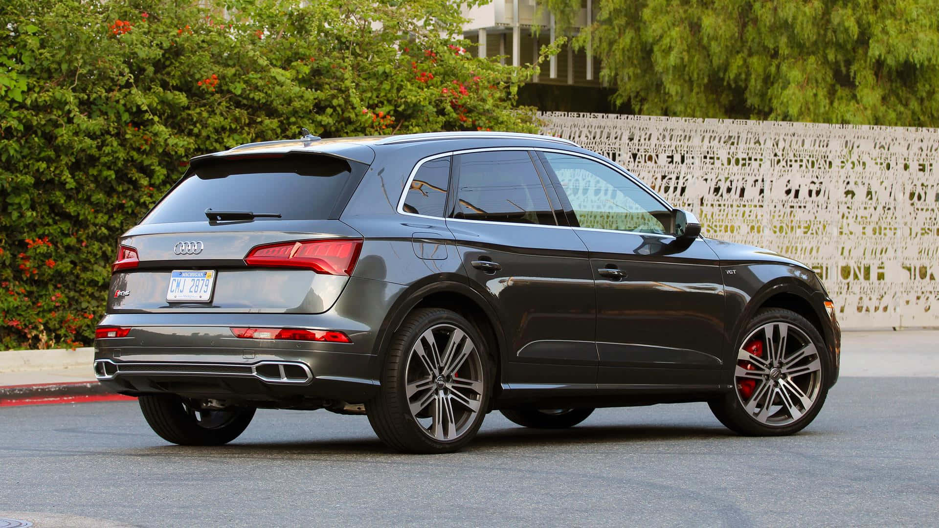 Audi Sq5 桌布 壁紙 — 一架型格灰色奧迪Q5 SUV停泊在瀝青路上，透過合金輪圈可見紅色制動卡鉗，四周環繞綠色灌木叢和白色裝飾圍欄。