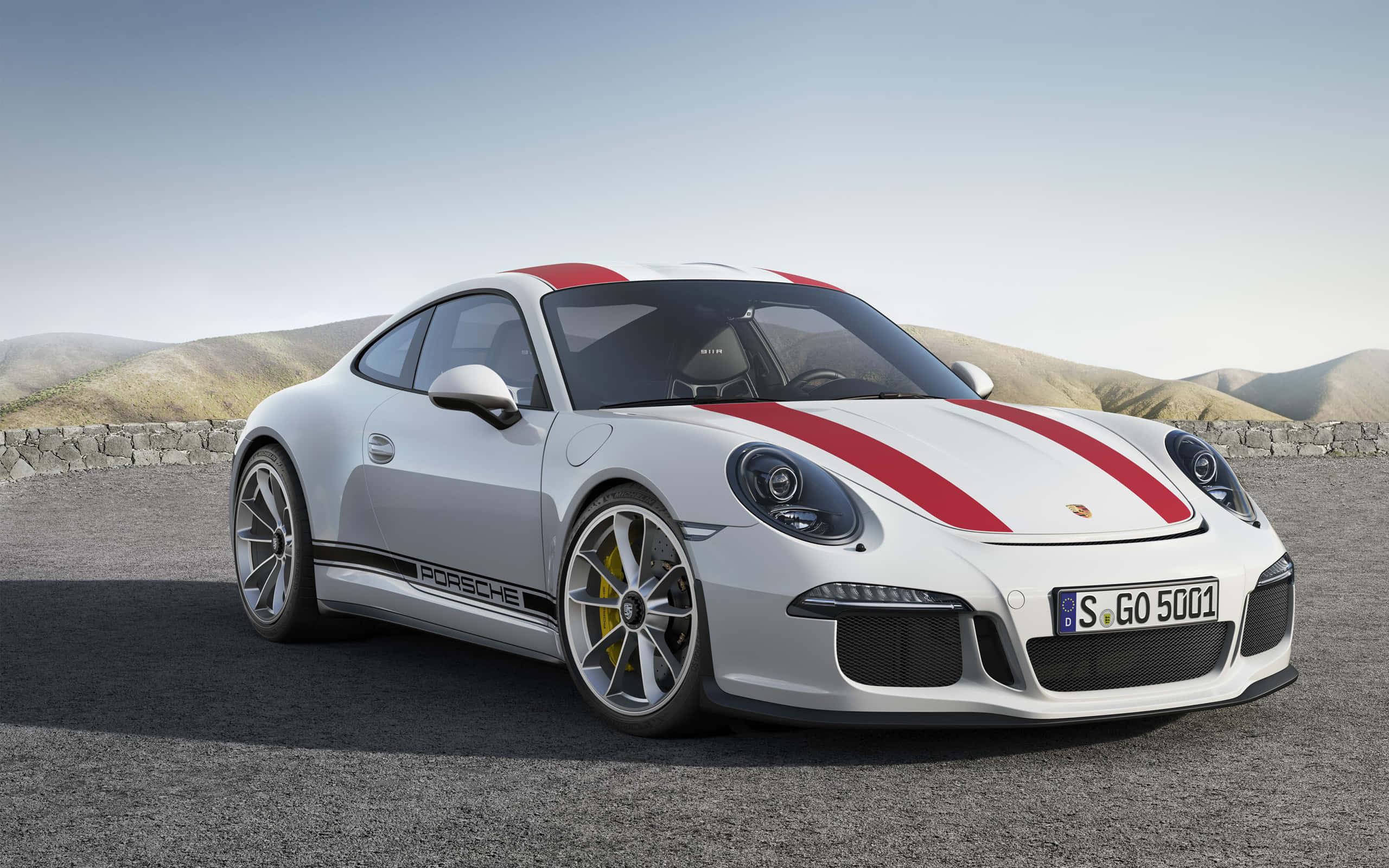 保時捷 桌布 壁紙 — 一架型格白色保時捷911 GT3 RS，帶有鮮明紅色賽車條紋，停泊在碎石路上，背景是連綿起伏的山丘，天空晴朗。
