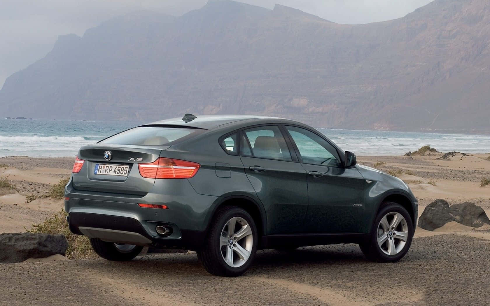 Bmw X6 桌布 壁紙 — 一架型格綠色BMW X6 SUV停泊在崎嶇的海岸沙灘上，背景有海浪拍打懸崖。