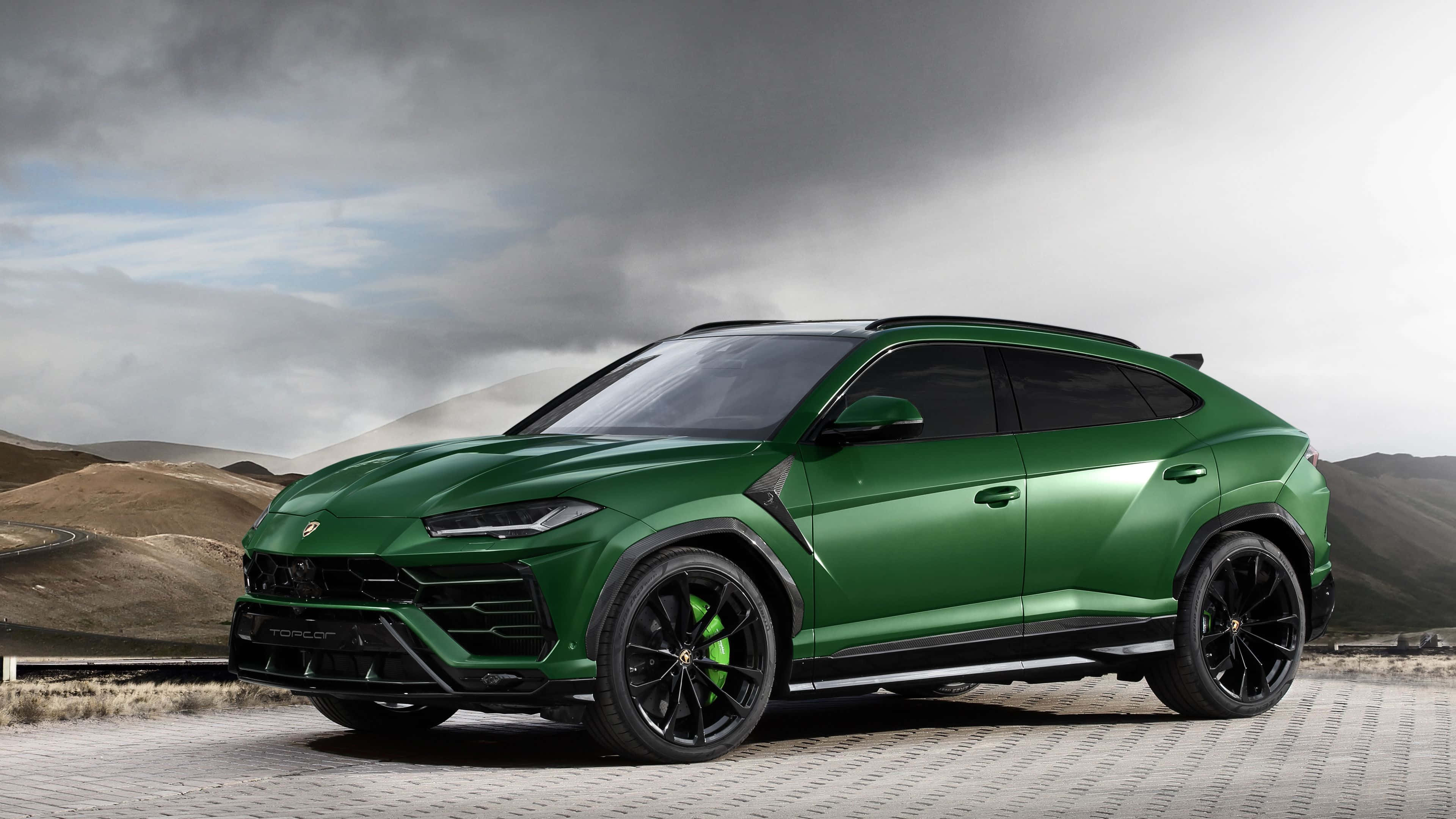 Lamborghini Urus 桌布 壁紙 — 一架型格綠色林寶堅尼 Urus SUV 停泊在鋪砌道路上，背景是戲劇性的多雲天空和連綿起伏的山丘。