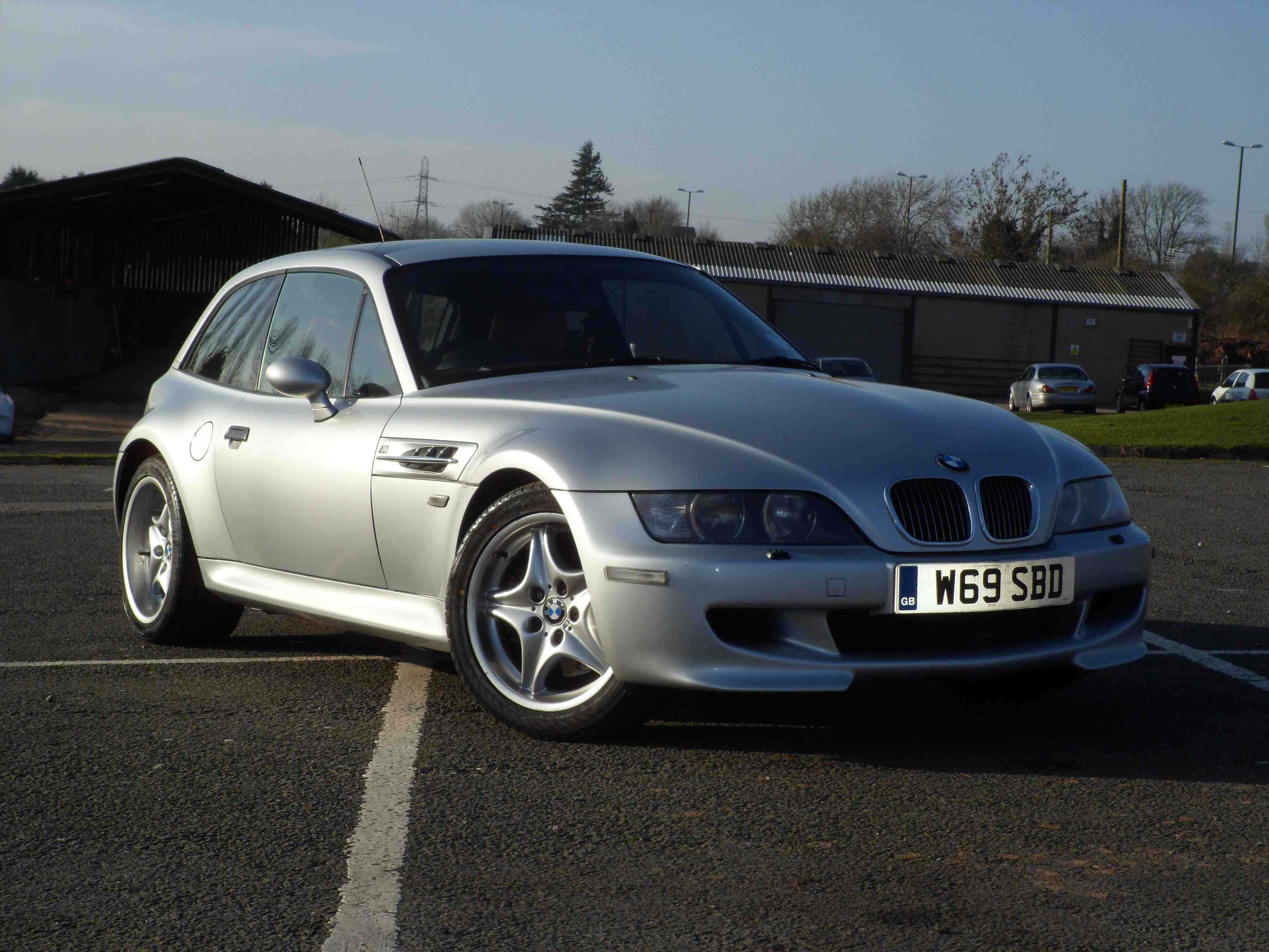 Bmw Z3 桌布 壁紙 — 一架型格銀色BMW Z3 M Coupe停泊在陽光普照的瀝青停車場，背景有工業建築物和光禿禿的樹木。