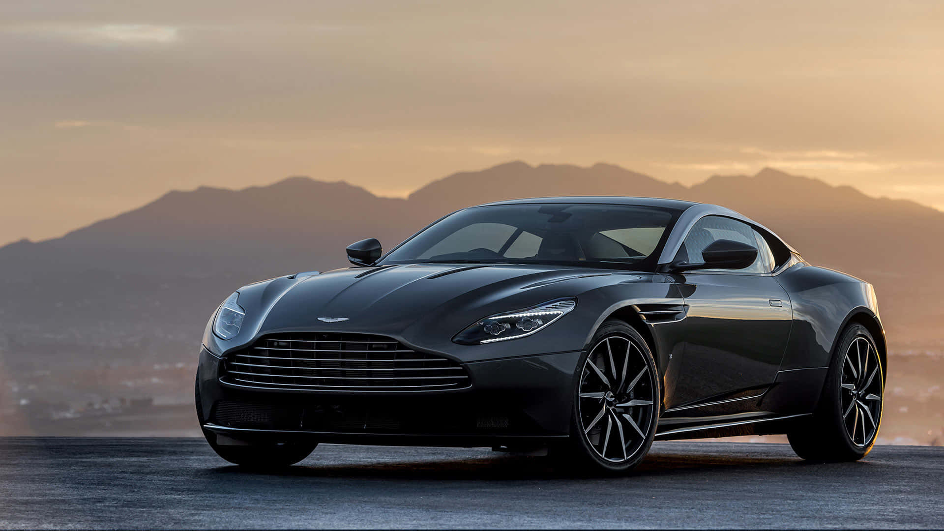 Aston Martin Db11 桌布 壁紙 — 一架型格黑色Aston Martin跑車在金色夕陽下閃耀，背景是遠山輪廓。