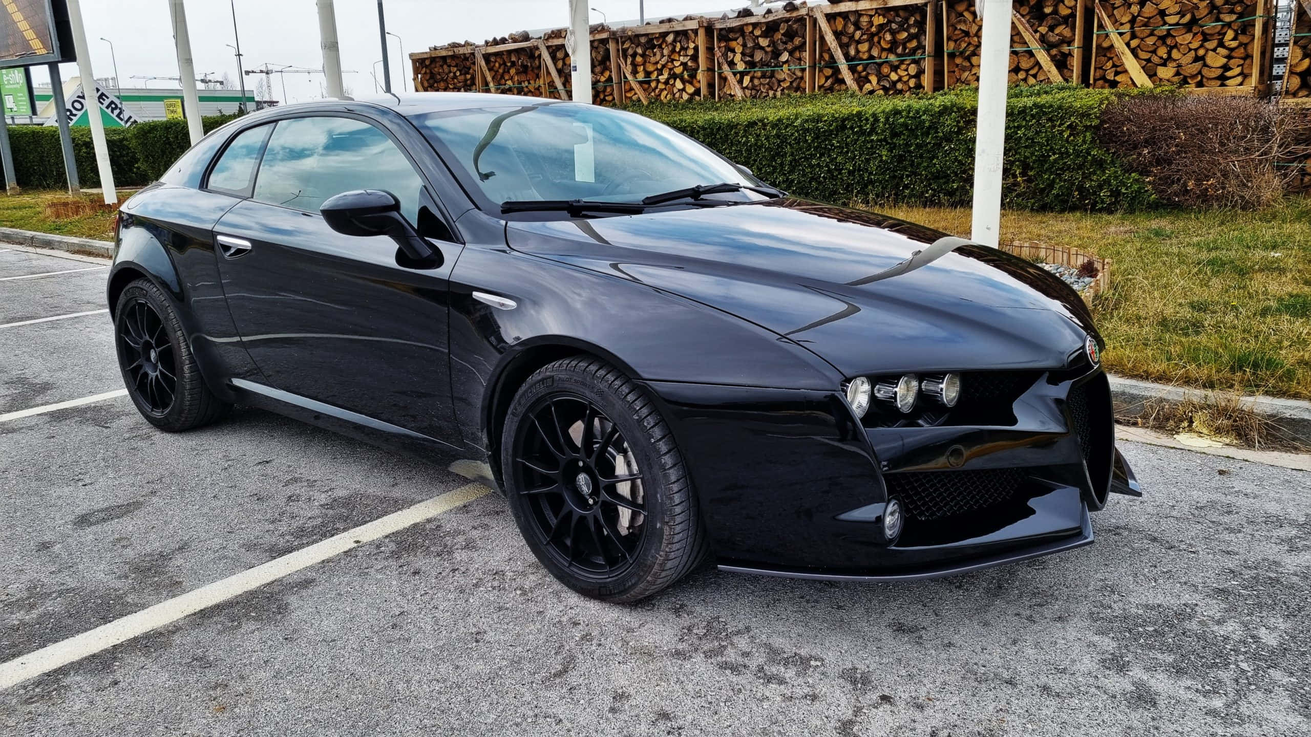 Alfa Romeo 159 桌布 壁紙 — 一架型格黑色愛快羅密歐跑車停泊在停車場，背景有堆疊木材和綠色籬笆。