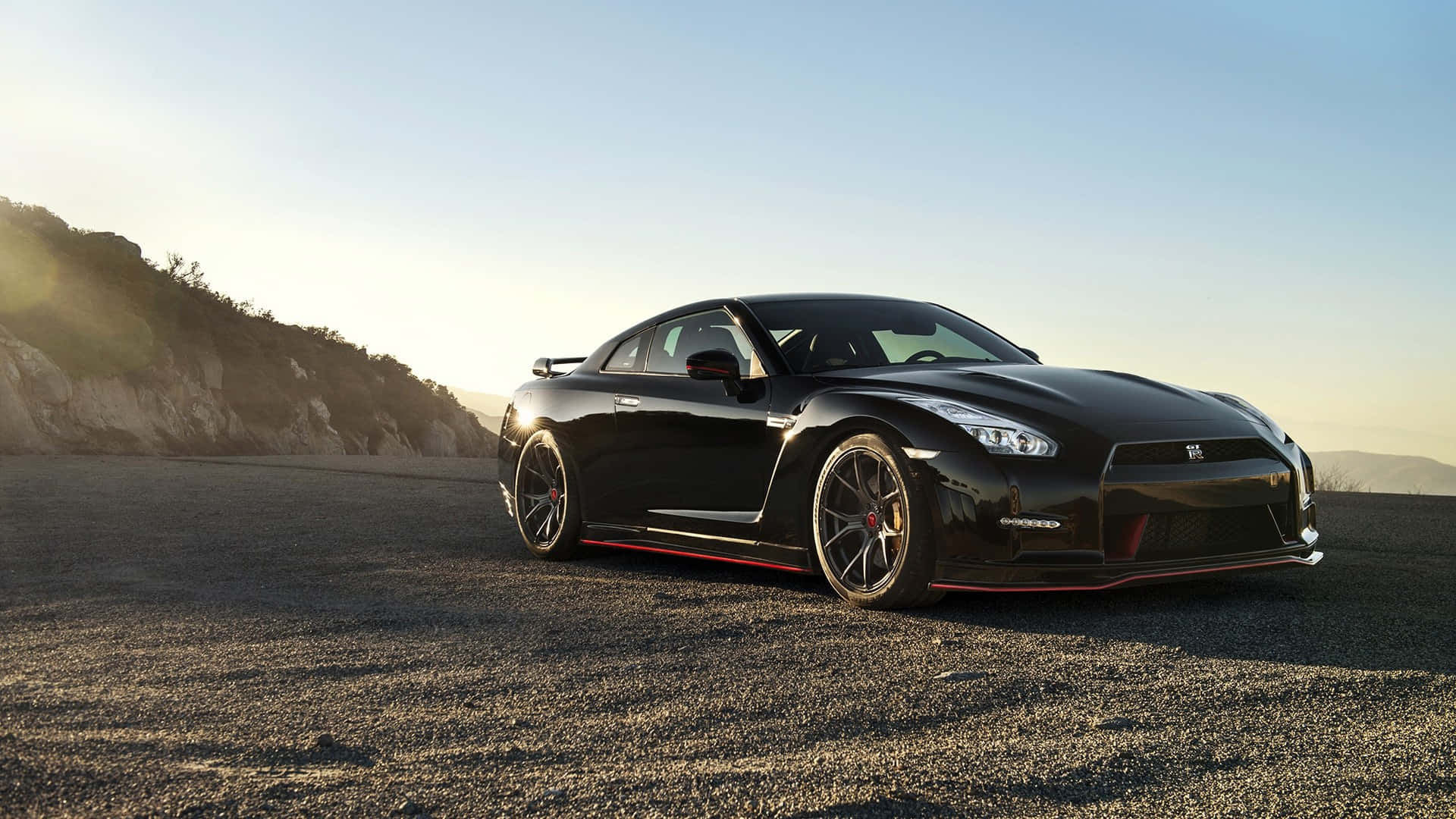 Gtr 桌布 壁紙 — 一架型格黑色日產GT-R跑車，帶有紅色點綴，停泊在日落時分的碎石路上，背景是山丘。