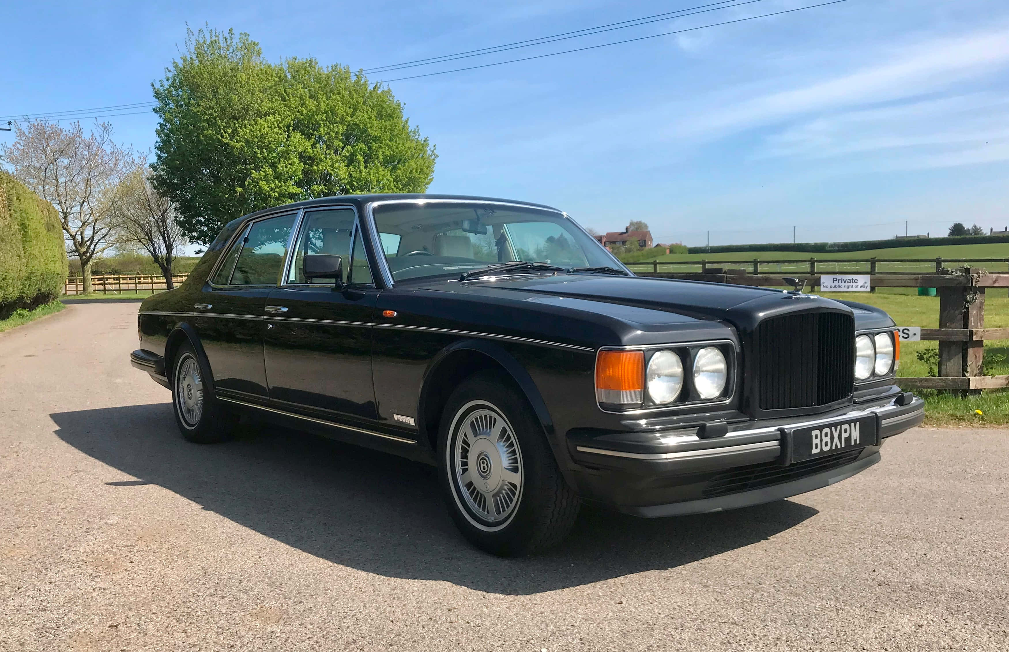 Bentley Eight 桌布 壁紙 — 一架型格黑色賓利房車在明亮陽光下閃閃發光，停泊在碎石車道上，旁邊是翠綠的田野和樹木。