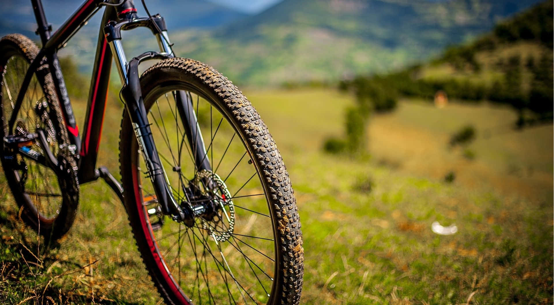 Mtb 桌布 壁紙 — 一架堅固嘅山地單車，車胎沾滿泥濘，停喺草地地形上，背景係起伏嘅綠色山丘同遠處嘅山脈。