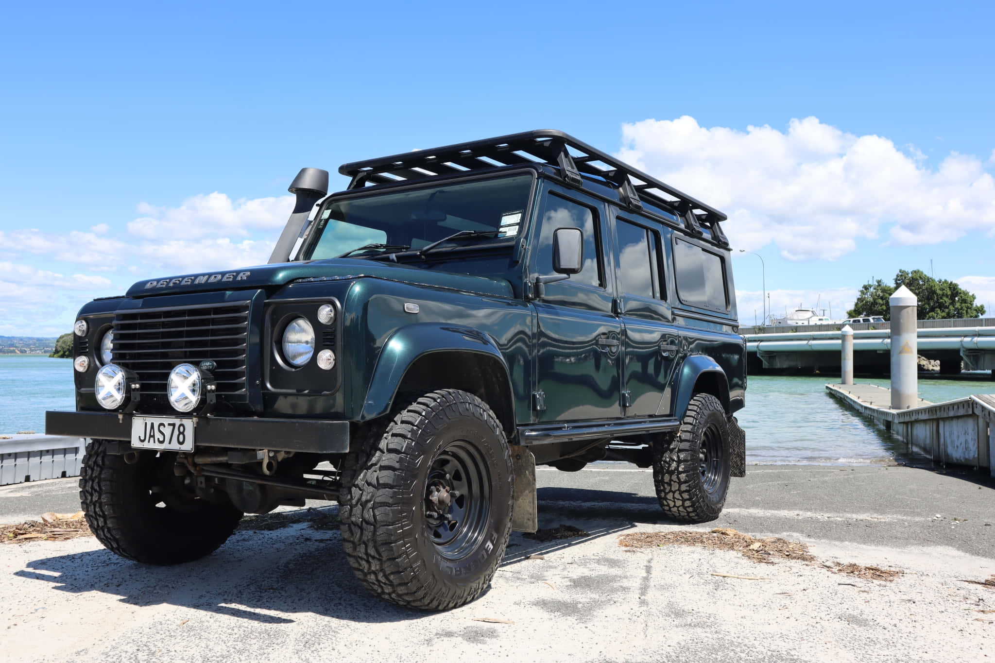 Land Rover Defender 桌布 壁紙 — 一架堅固的深綠色 Land Rover Defender，配備車頂架和超大輪胎，停泊在混凝土碼頭上，旁邊是平靜的水面，上方是藍天白雲。